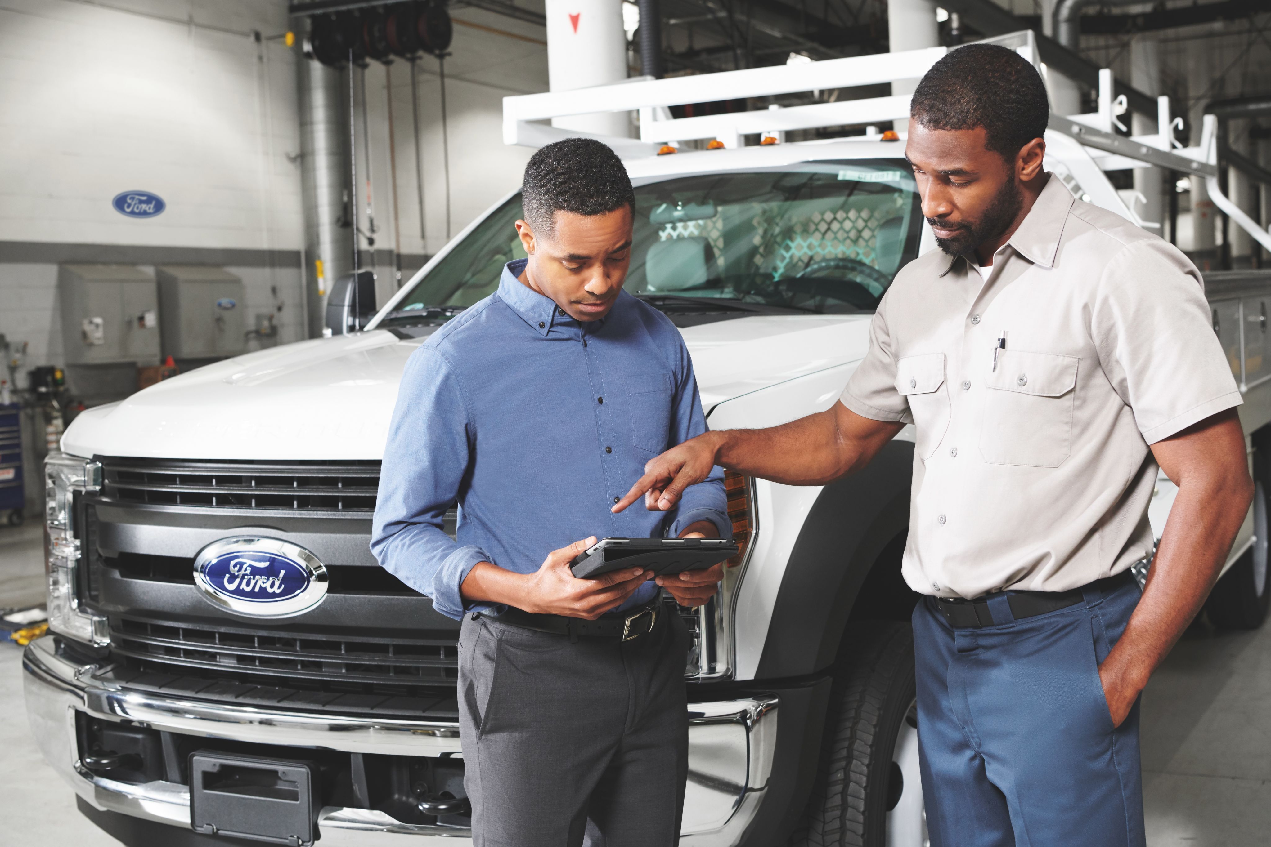 Two men look at a tablet with a Ford vehicle behind them