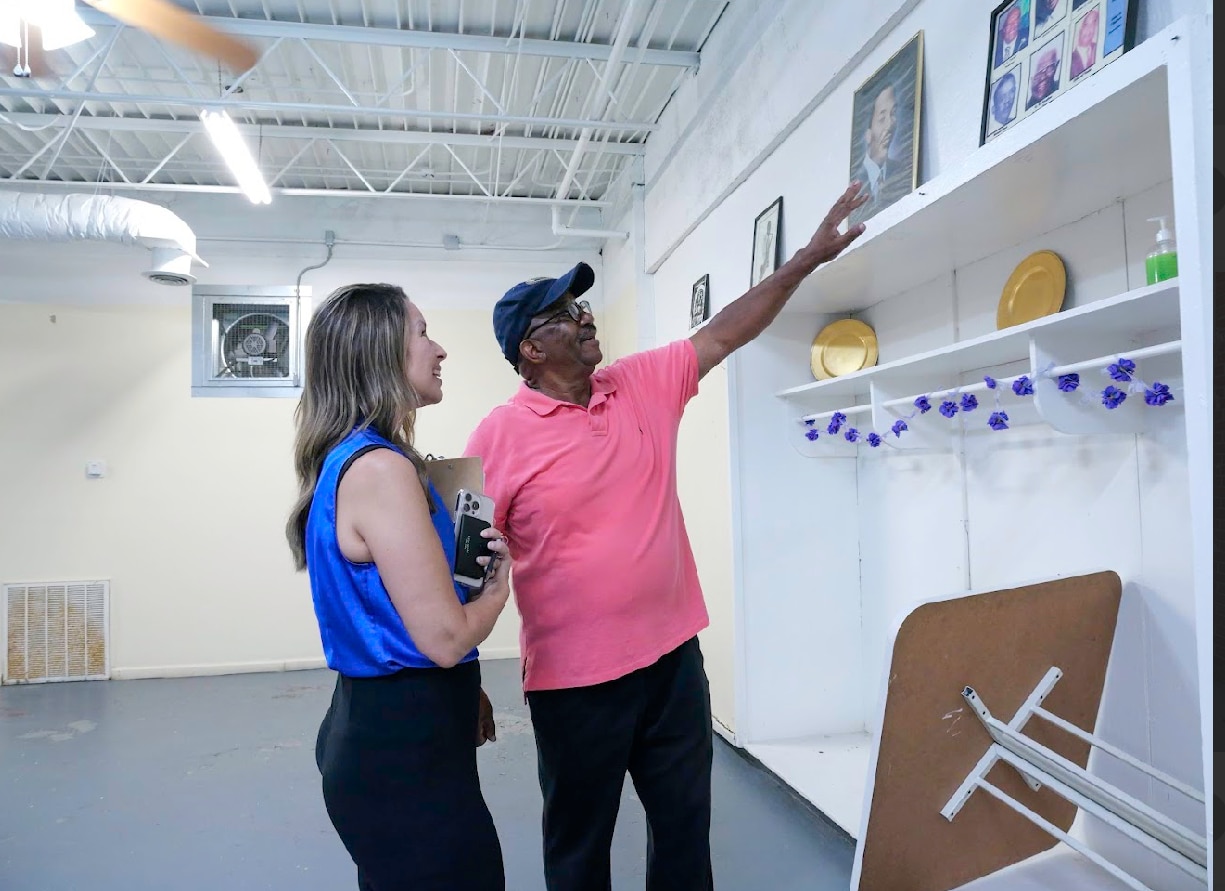 A man and woman stand inside of a building that looks to be in the middle of renovations. The man shows the woman a portrait on the wall.