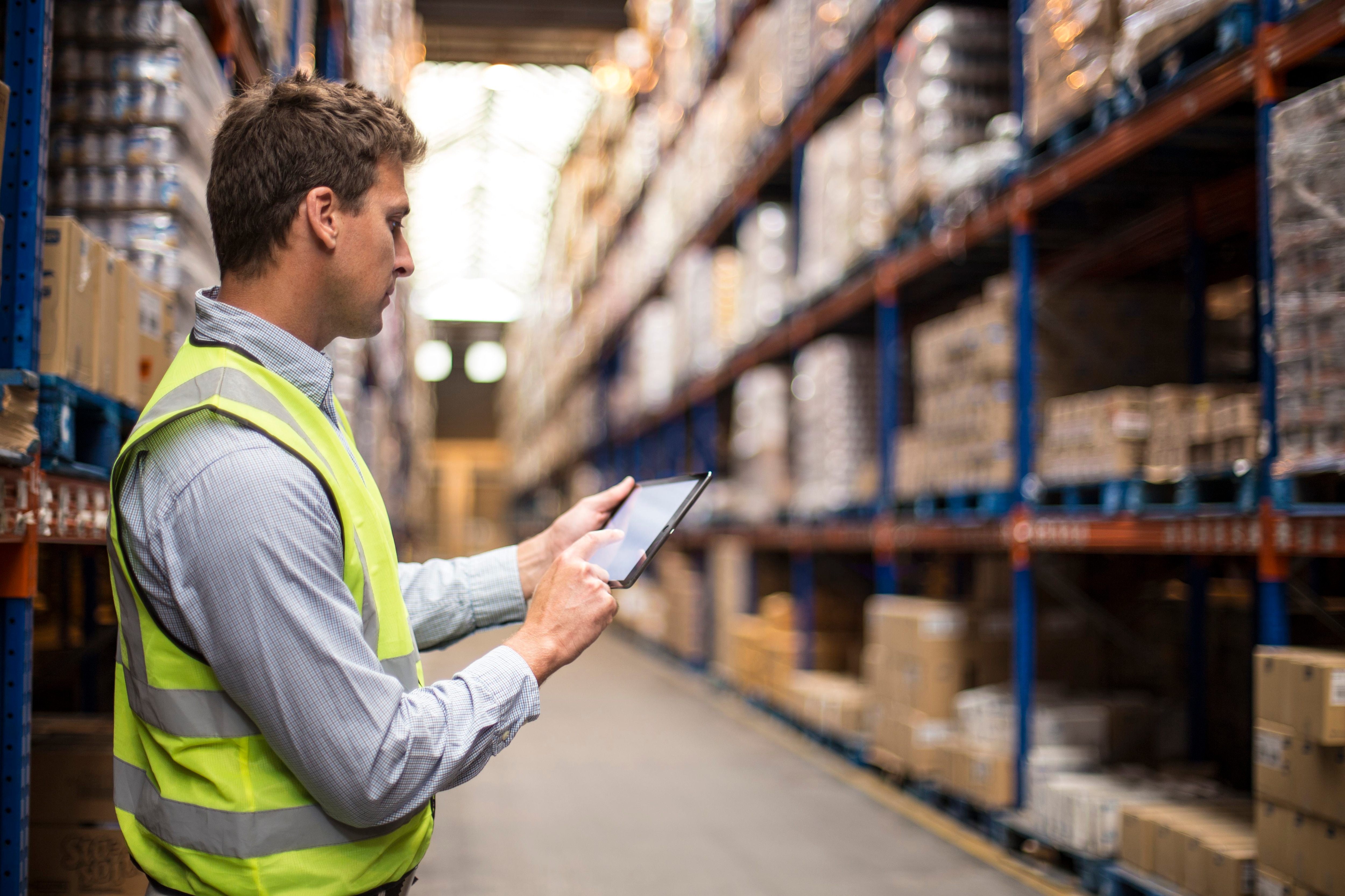 A man wearing a yellow vest stands in a warehouse with shelves full of boxes and looks at a tablet.