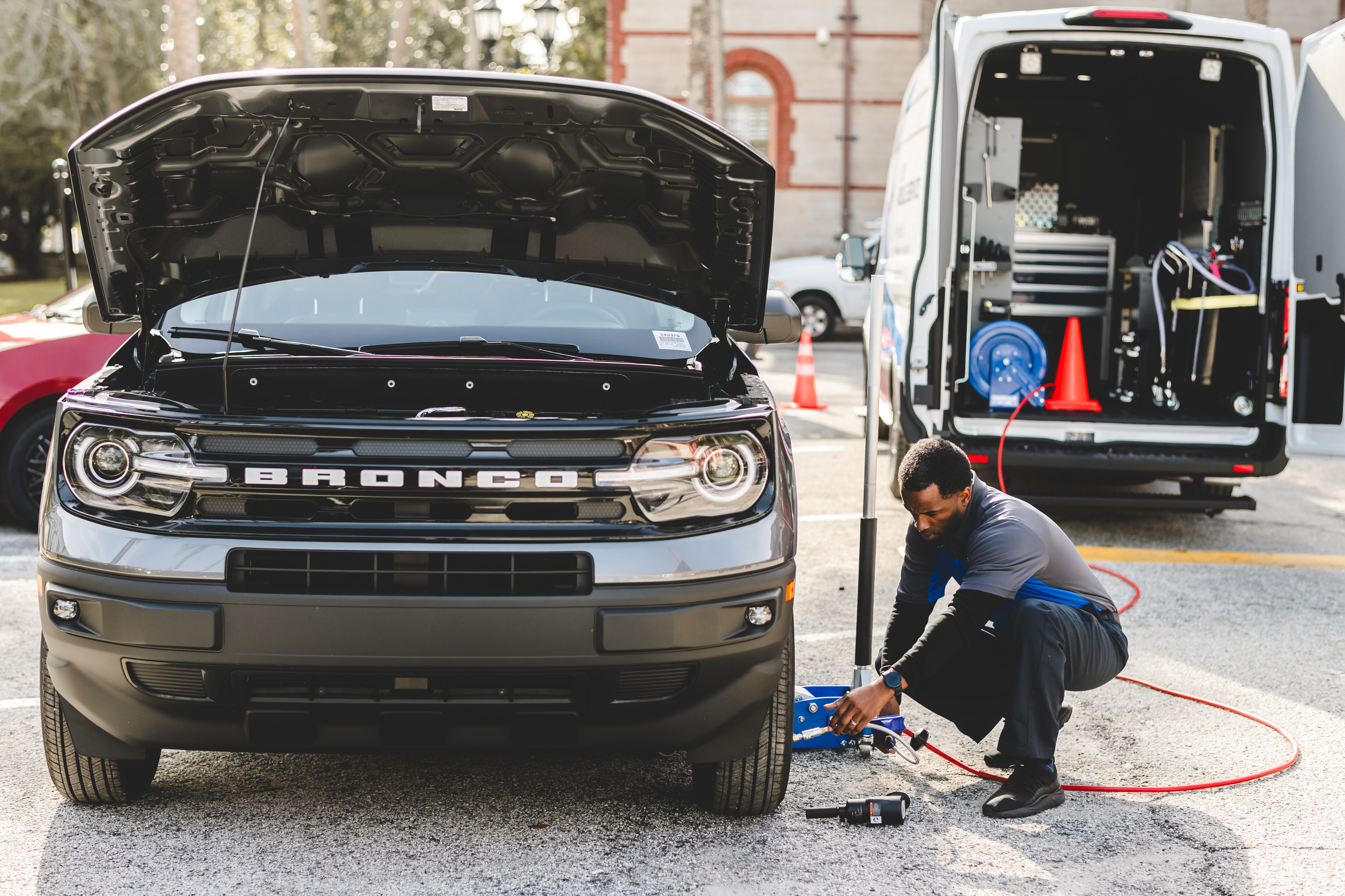 A man leans down to work on the tire of a ford bronco. In the background, a Ford Transit Mobile Service van is parked with its back doors open.