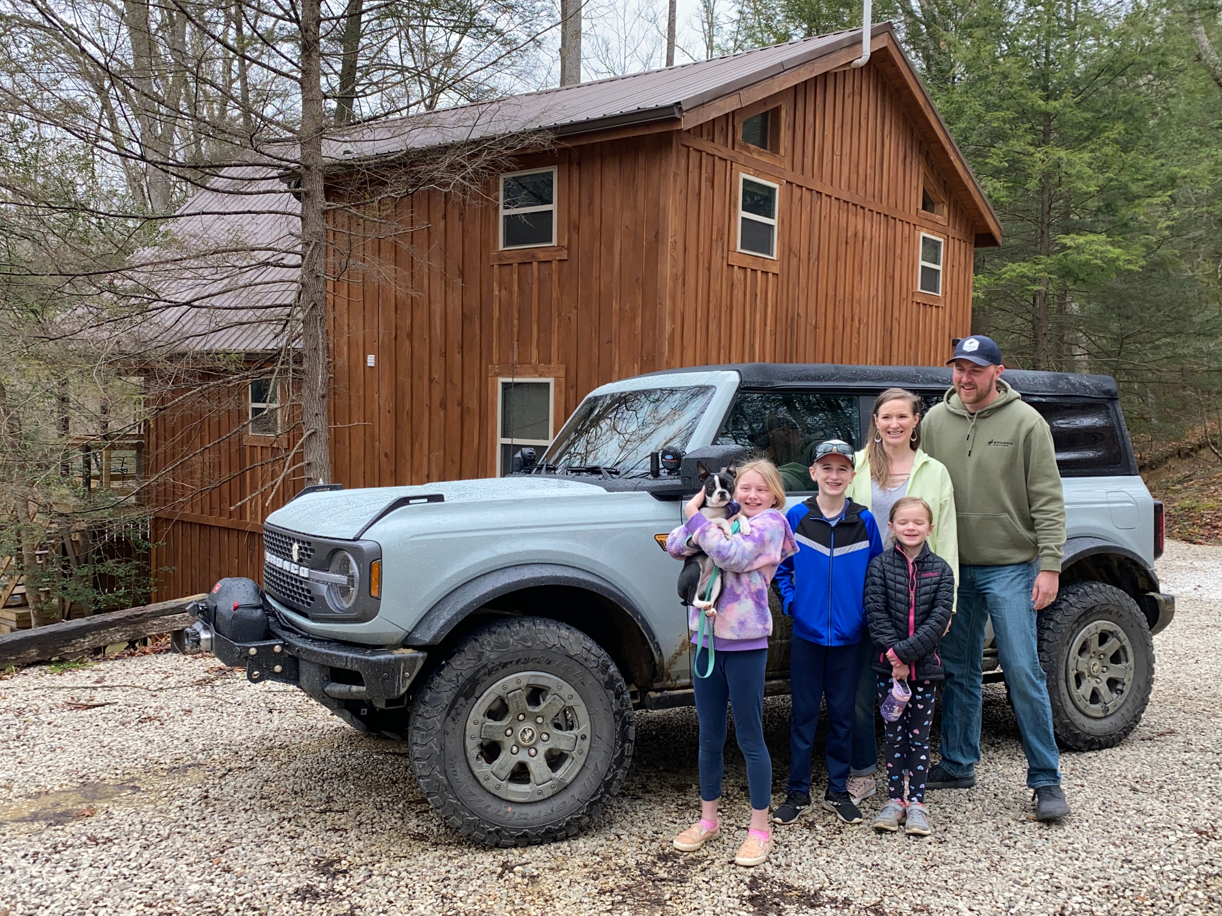 A family poses in front of a Ford Bronco. A rustic-looking, large, wooden cabin and trees are in the background. A girl in the photo holds a young dog.