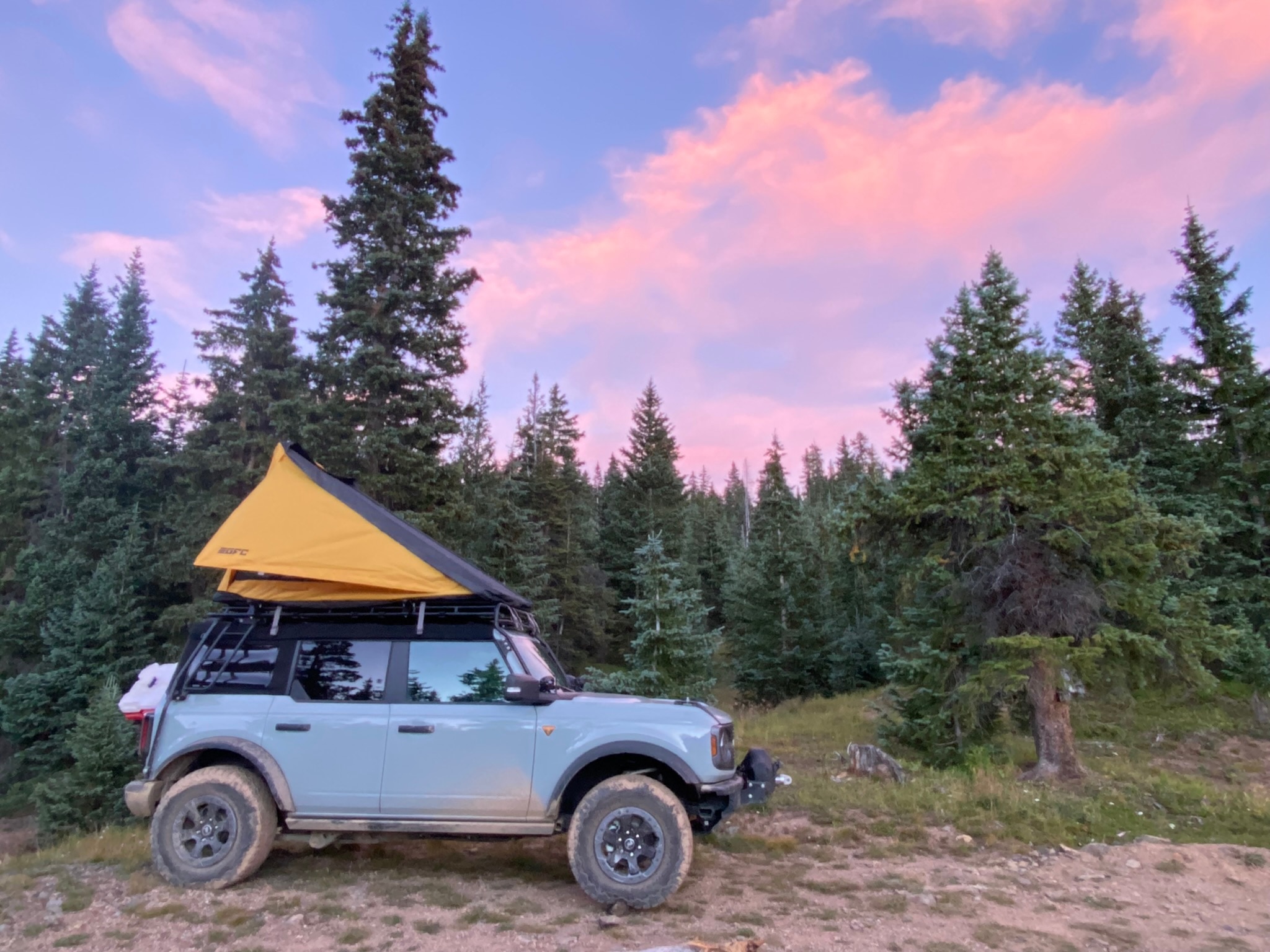 A blue Bronco with a tent on top is parked in the woods. The backdrop is of a forest and a purply sky at sunset with bright pink clouds,