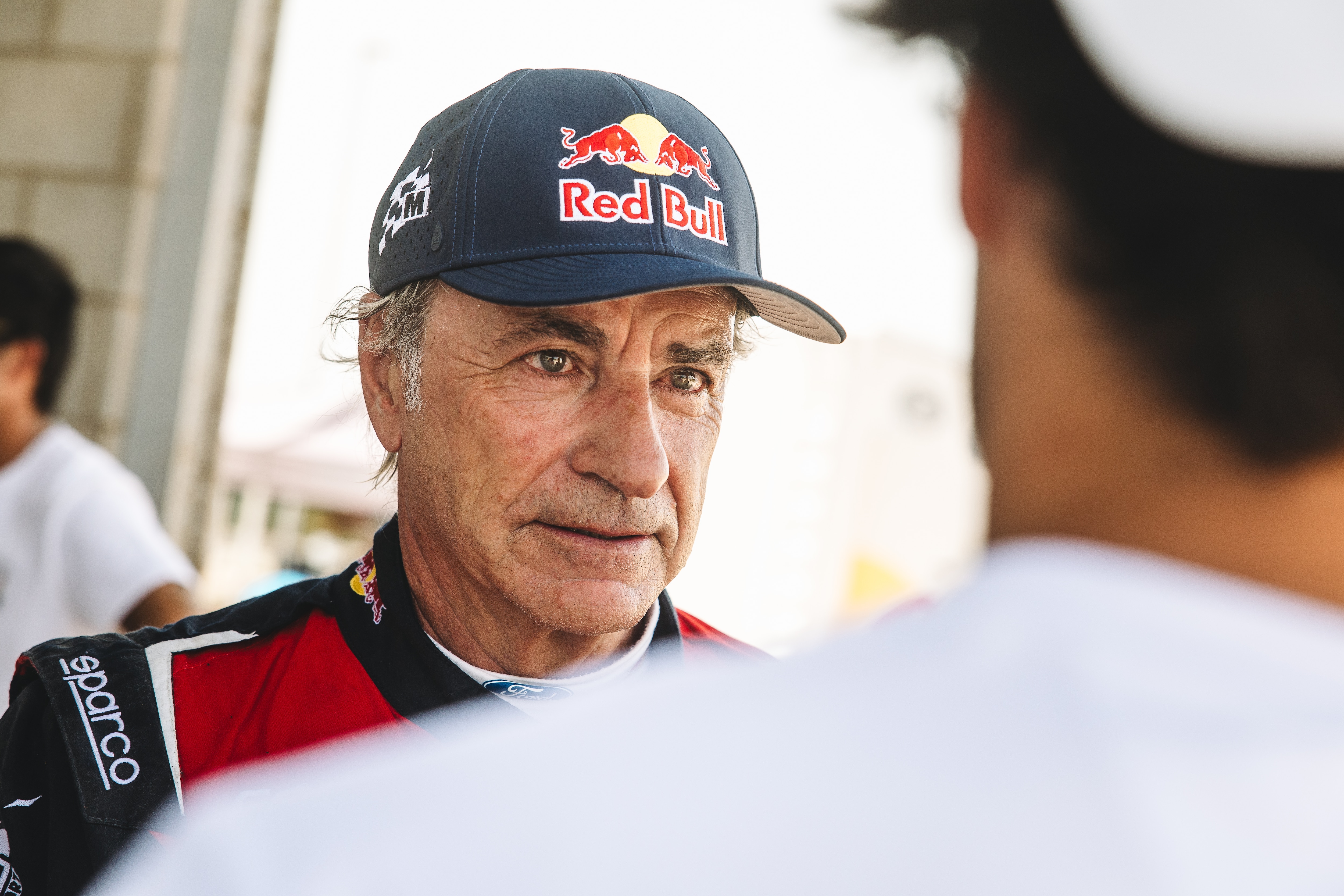 Carlos Sainz Senior wearing a black hat with a Red Bull logo and looking toward the camera over the shoulder of his son, who is wearing a white shirt and hat and has his back toward the camera