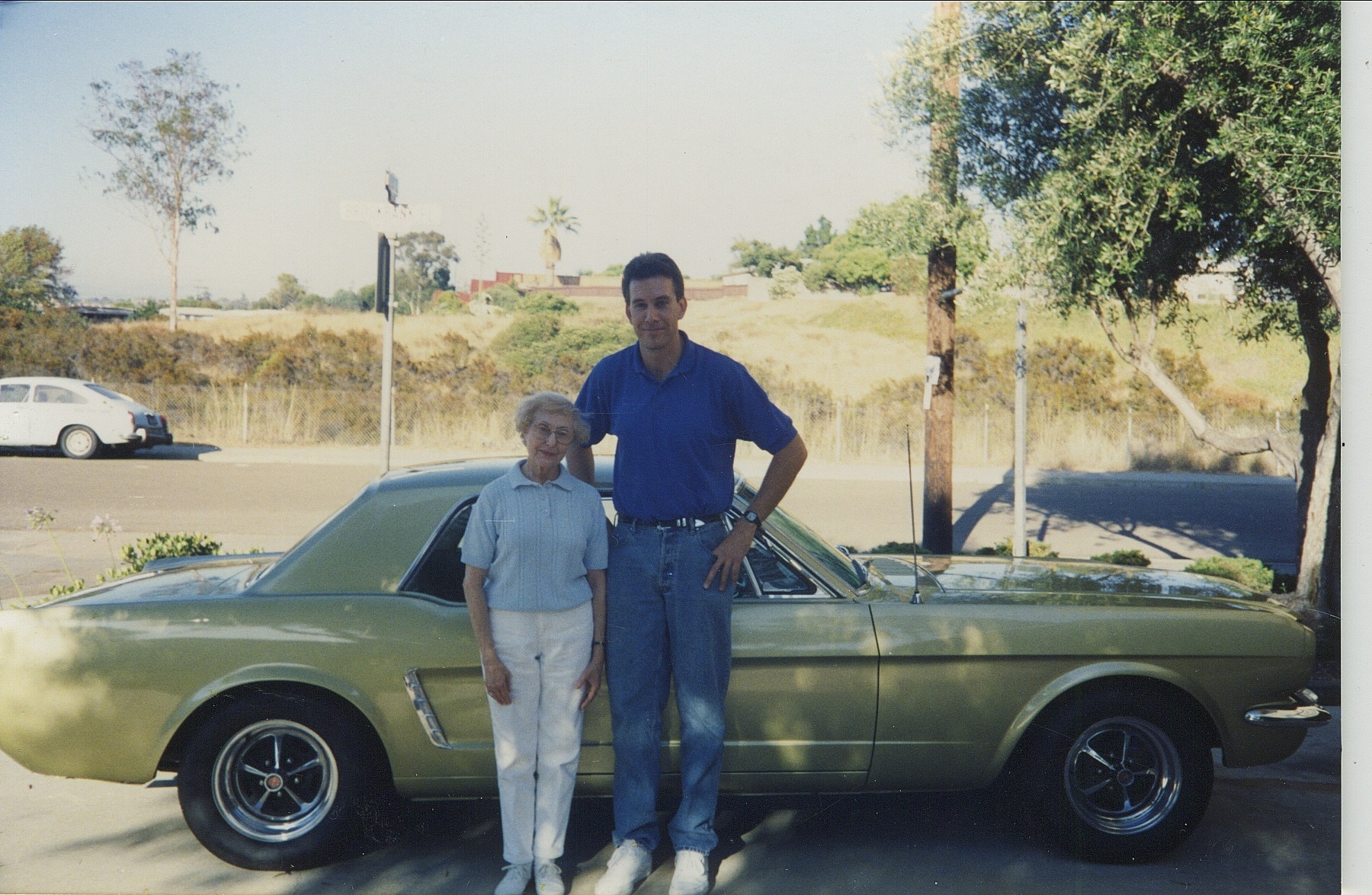 An older woman and a young man stand in front of a Mustang in the 1990s.