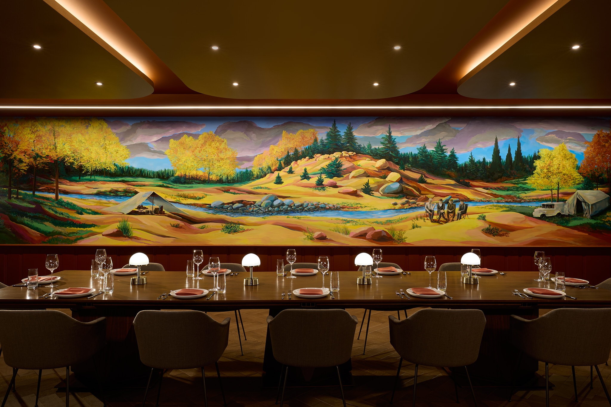 A colorful mural of a mountainous landscape with a tent in the foreground is pictured behind a long dining table in a bar in the newly renovated Dearborn Inn
