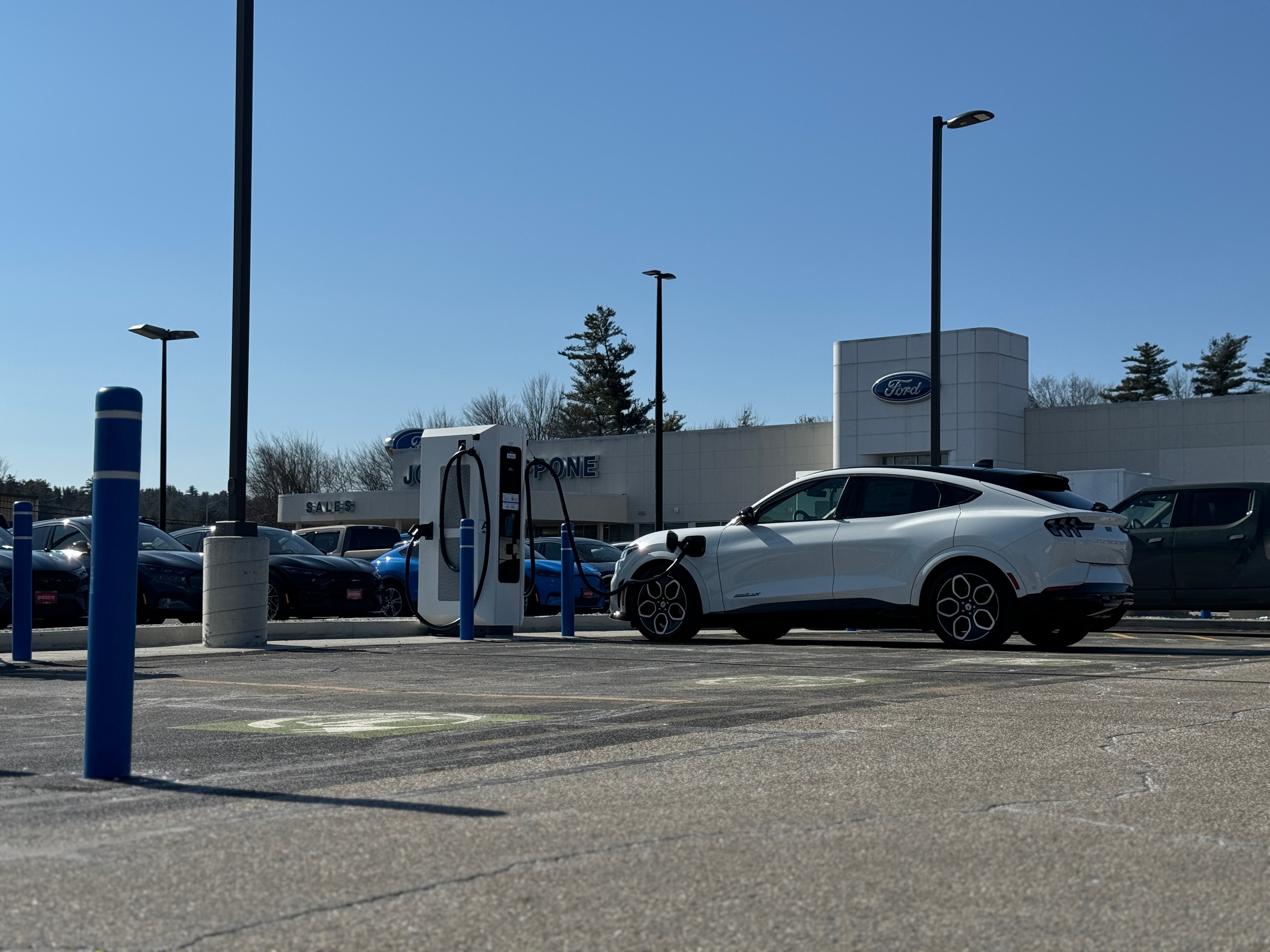 A white Ford Mustang Mach-E charges on a new charger in a Ford dealership parking lot.