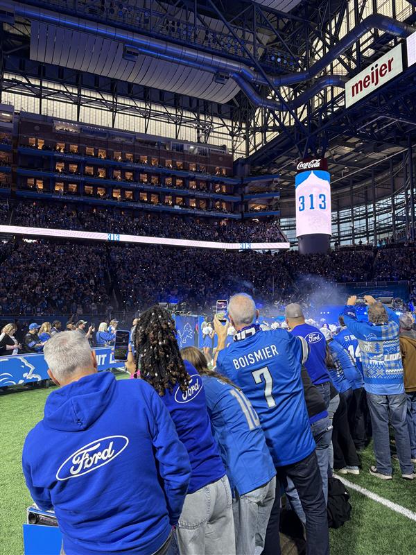 A group of people in Ford attire stand on the Detroit Lions field and film the pre-game festivities with their phones.