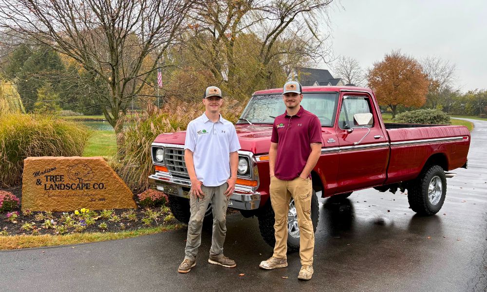 Two men stand near a vintage red Ford truck and a stone sign that reads "Madison Tree & Landscape Co."