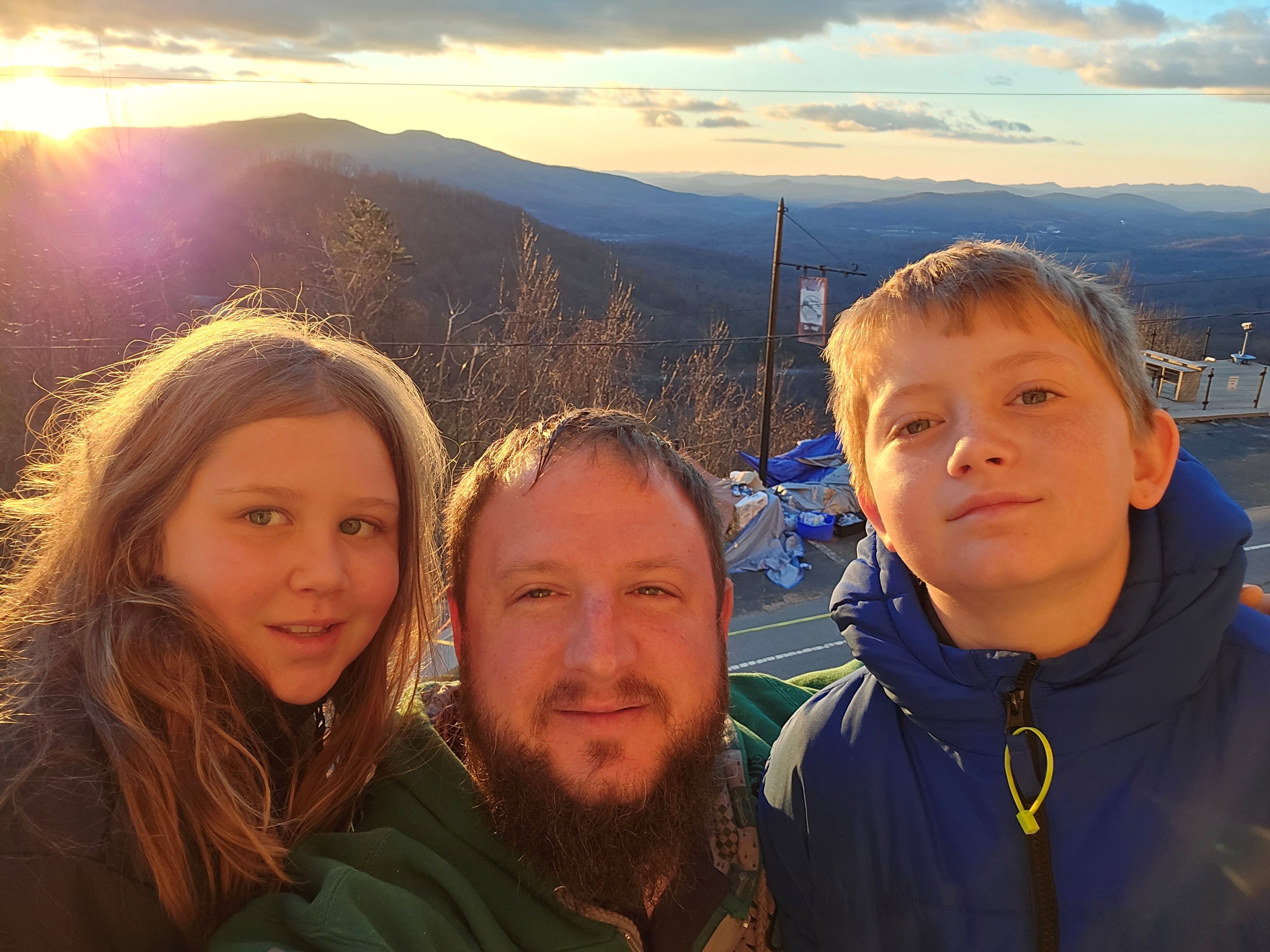 A man takes a selfie with his kids in front of a mountain sunset.