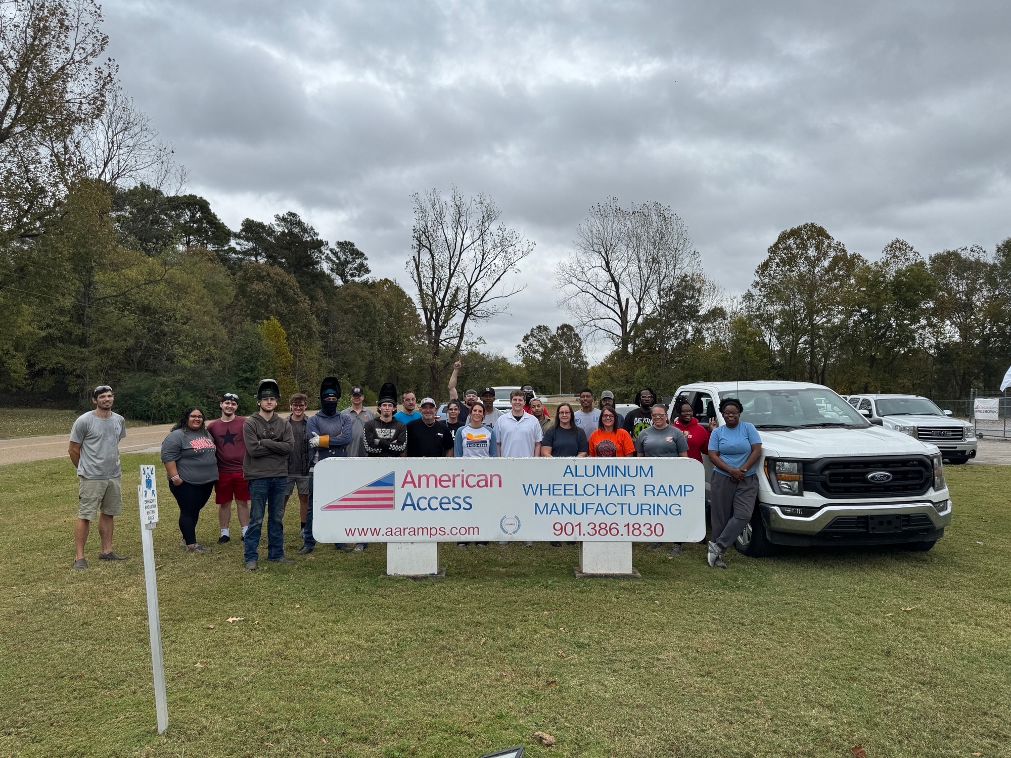 A group of people stand near a white truck and hold a sign that reads "American Access - Aluminum Wheelchair Ramp Manufacturing"