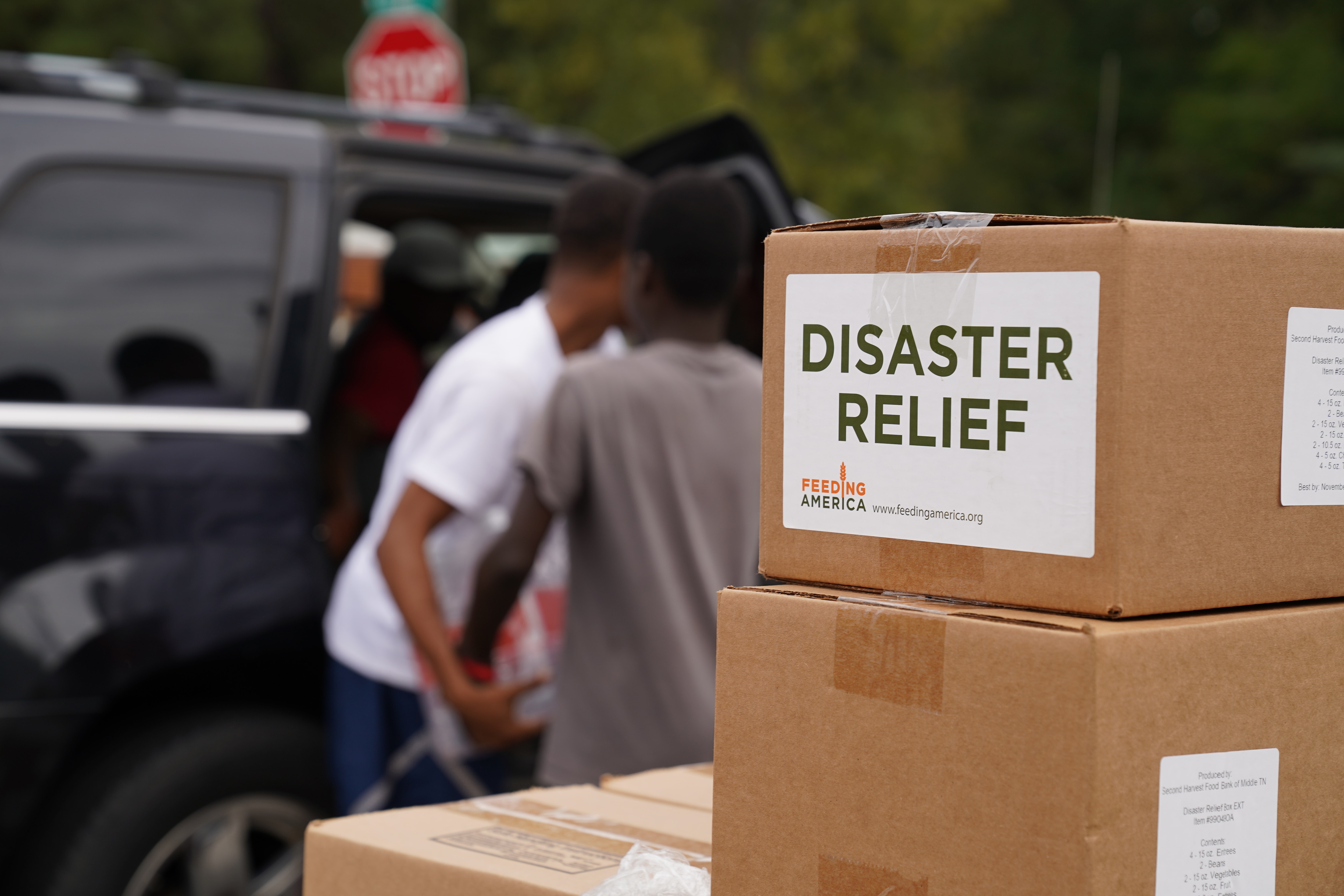 In the foreground, a stack of boxes marked "disaster relief" can be seen. Two men load items into a car in the background.