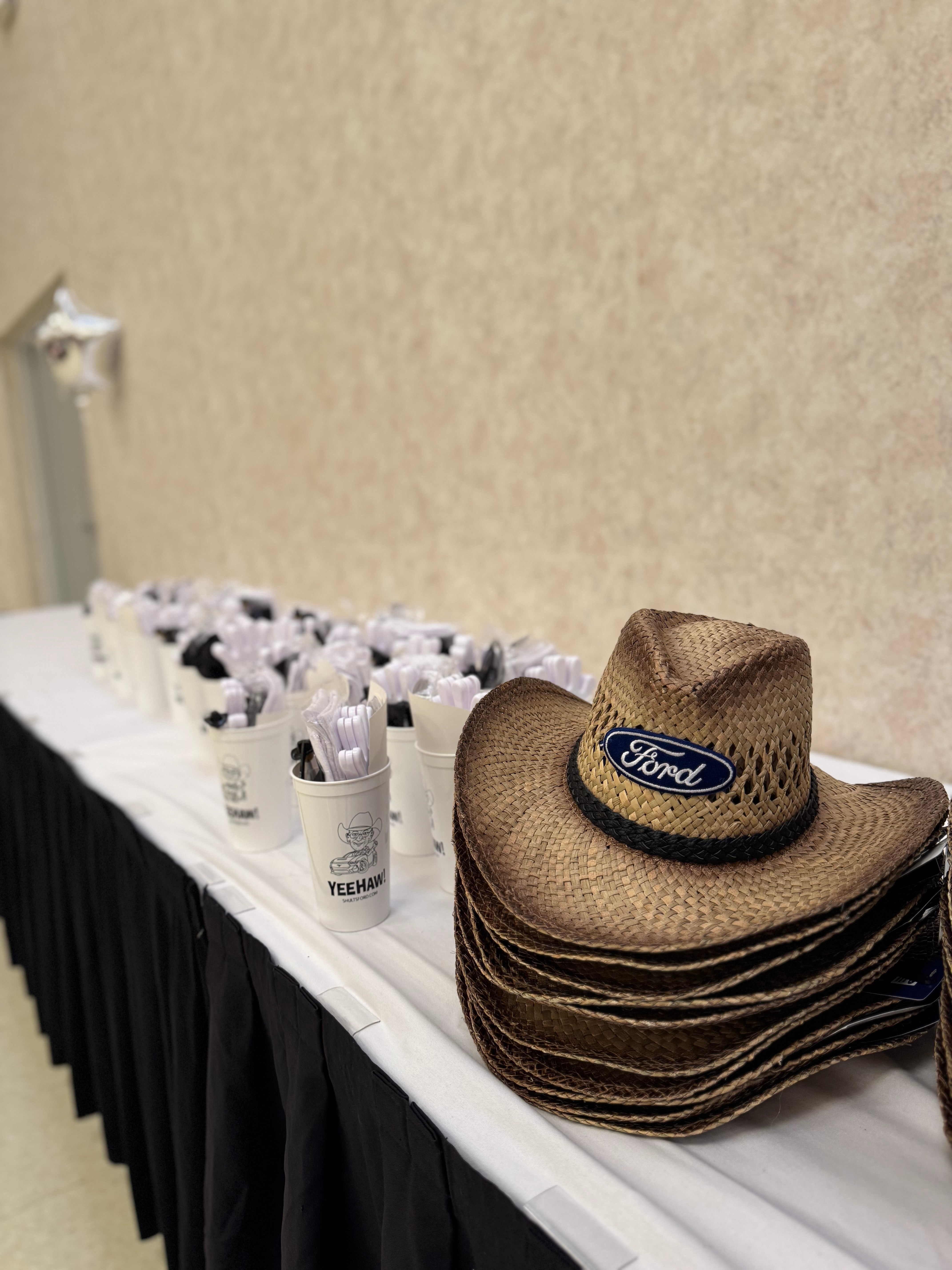 Straw hats with the Ford logo on the front sit on a table next to white cups that say "Yeehaw" 
