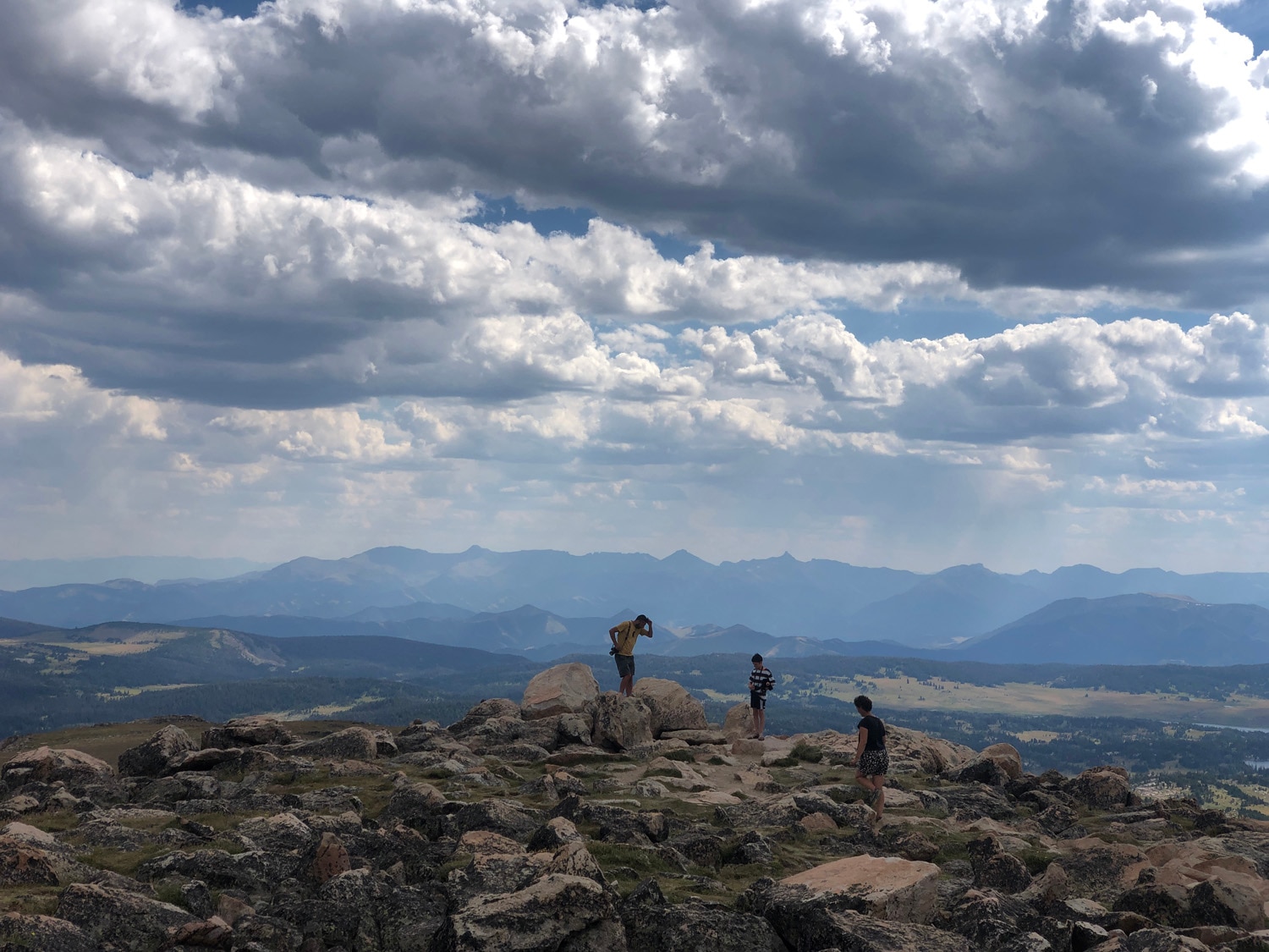 Three people stand atop a hilly peak, close to the clouds, with vast mountainous landscapes behind them.