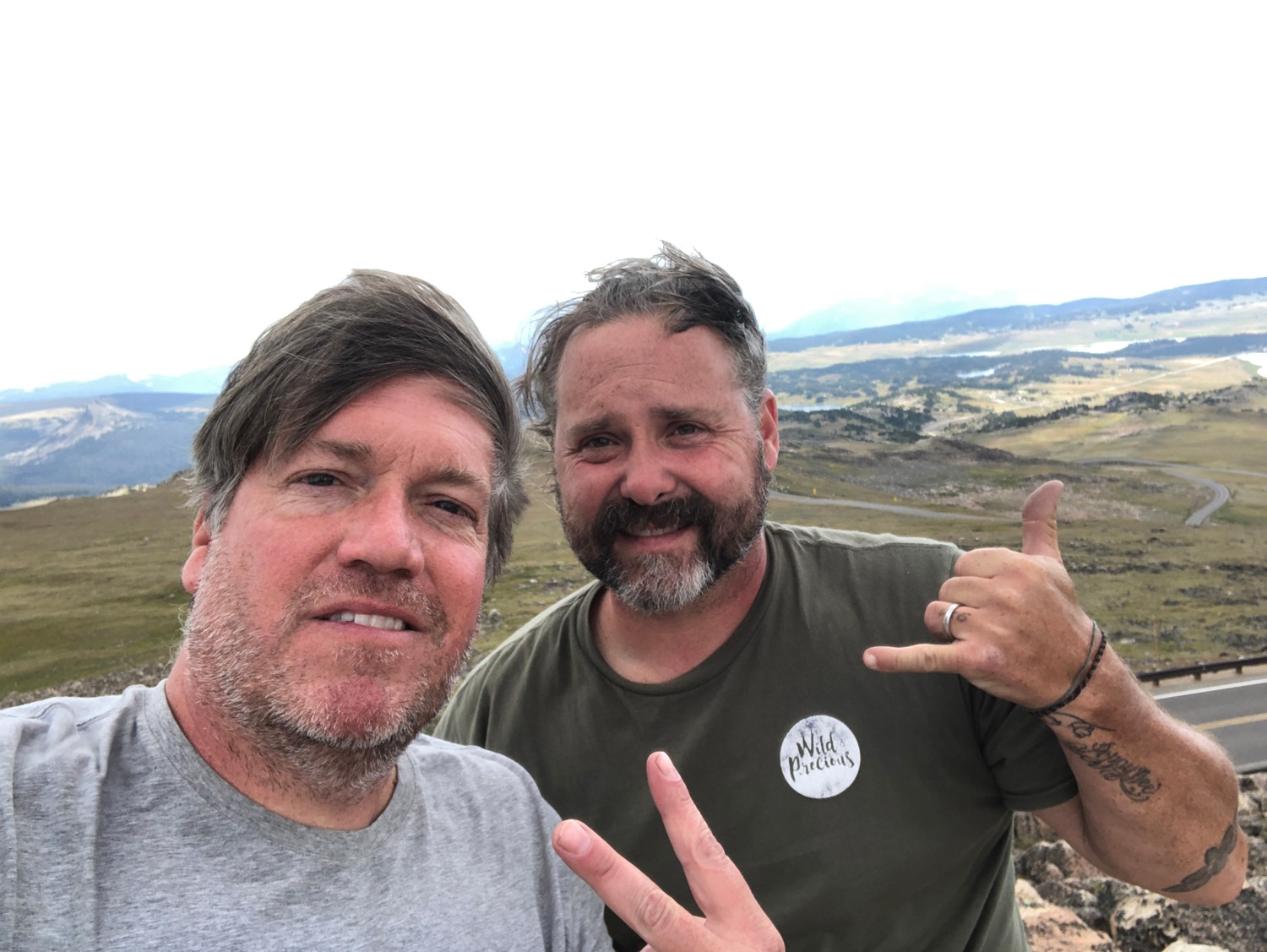 Two men smile for a selfie together with a rolling green landscape in the background.