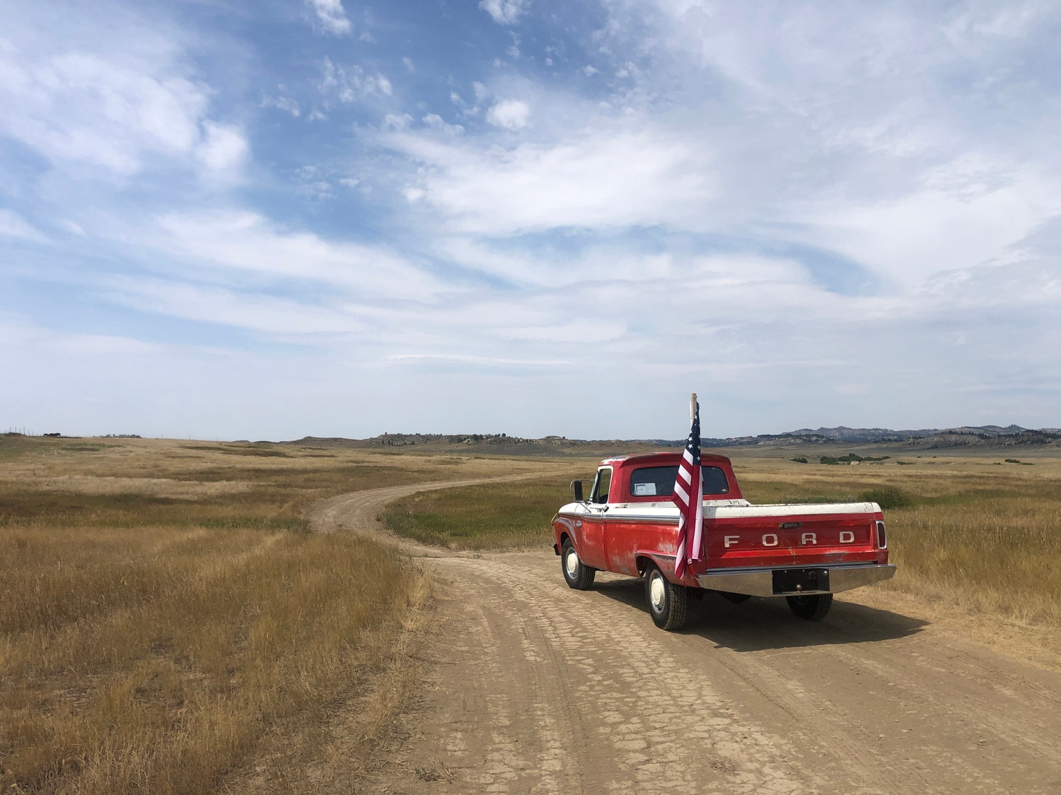 A red vintage Ford truck drives down a dirt road, flying an American flag off the back, with a blue sky above filled with wispy clouds.