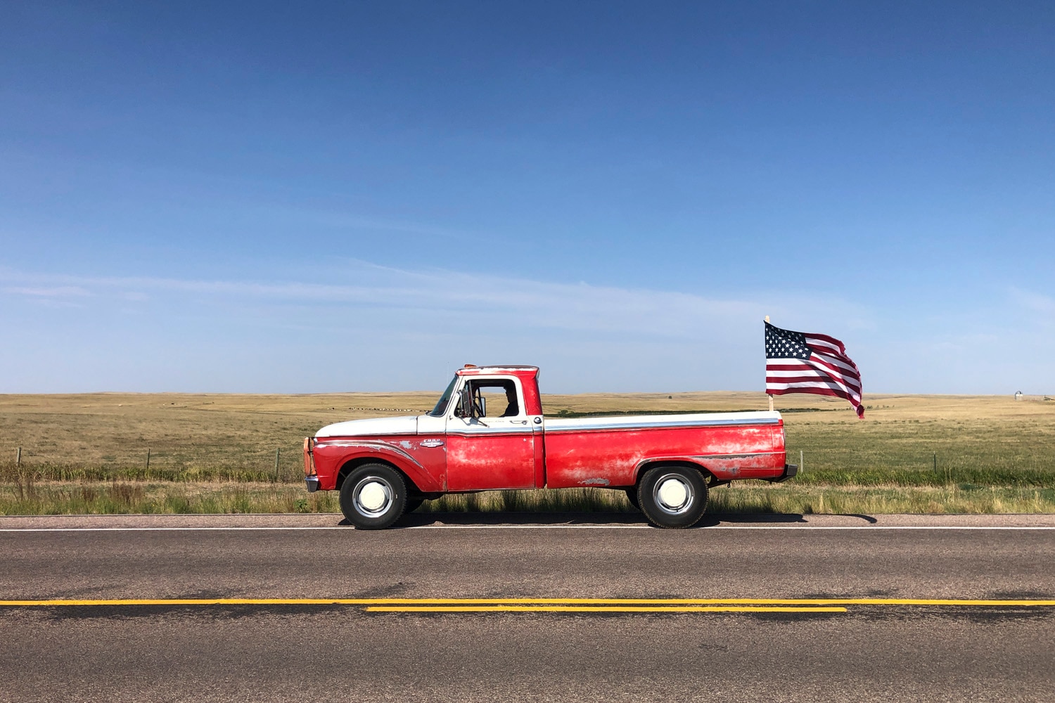 A red vintage Ford truck drives down a dirt road, flying an American flag off the back, with a blue sky above filled with wispy clouds.