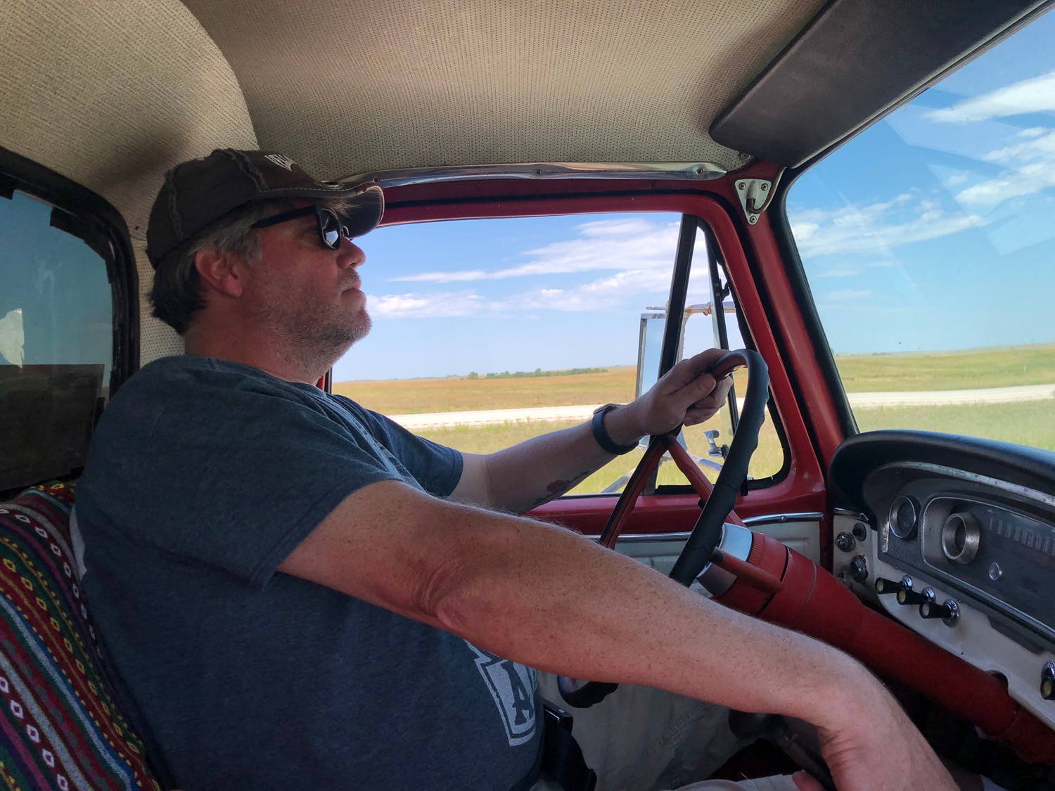 Viewed from the passenger seat, a man in a hat and sunglasses drives a vintage truck. Out of his window, vast plains and blue sky.