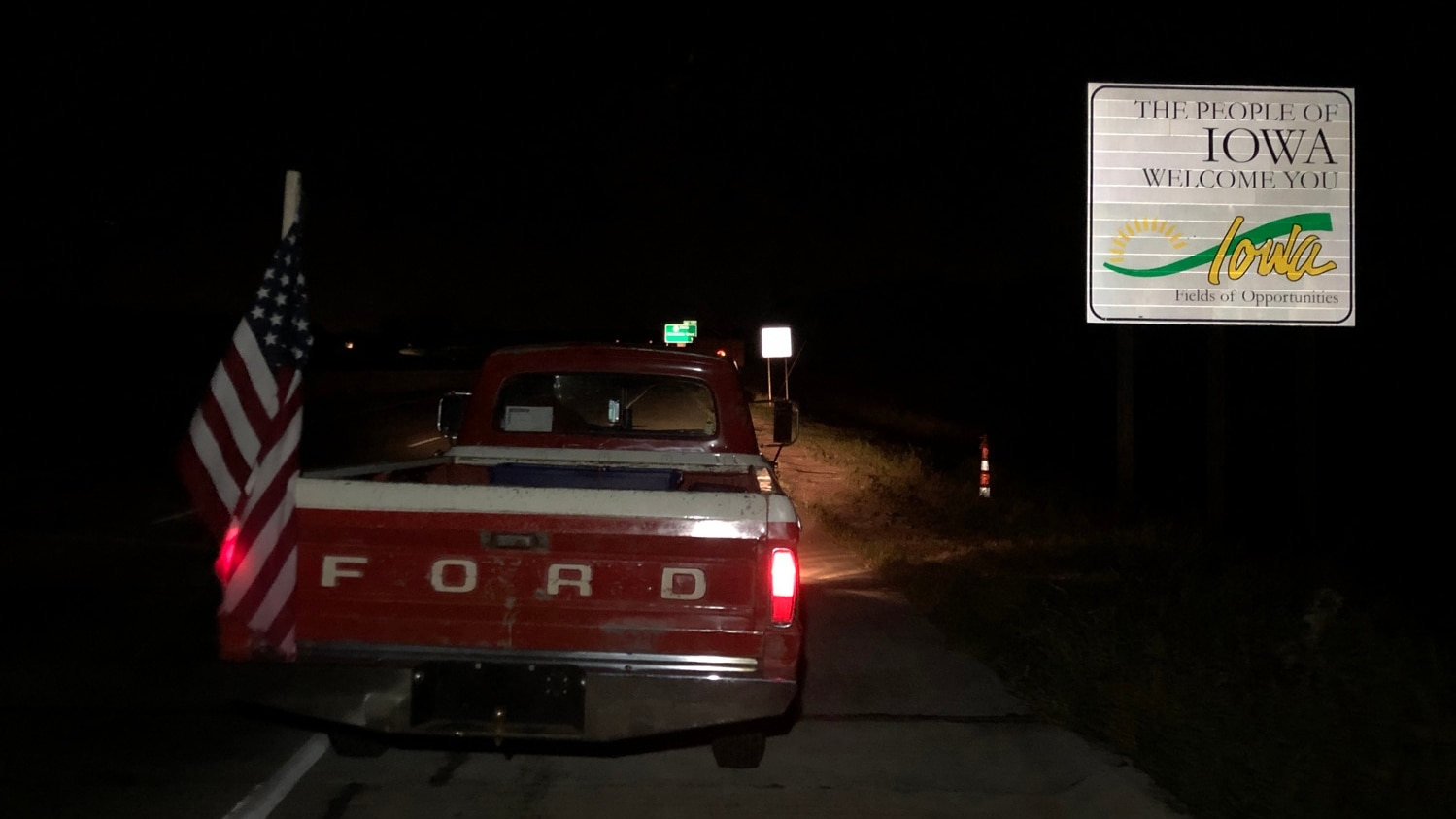 At night, seen from the back, a vintage Rod truck with an American flag attached to the bed is parked on the road next to a road sign that reads "The People of Iowa Welcome You"