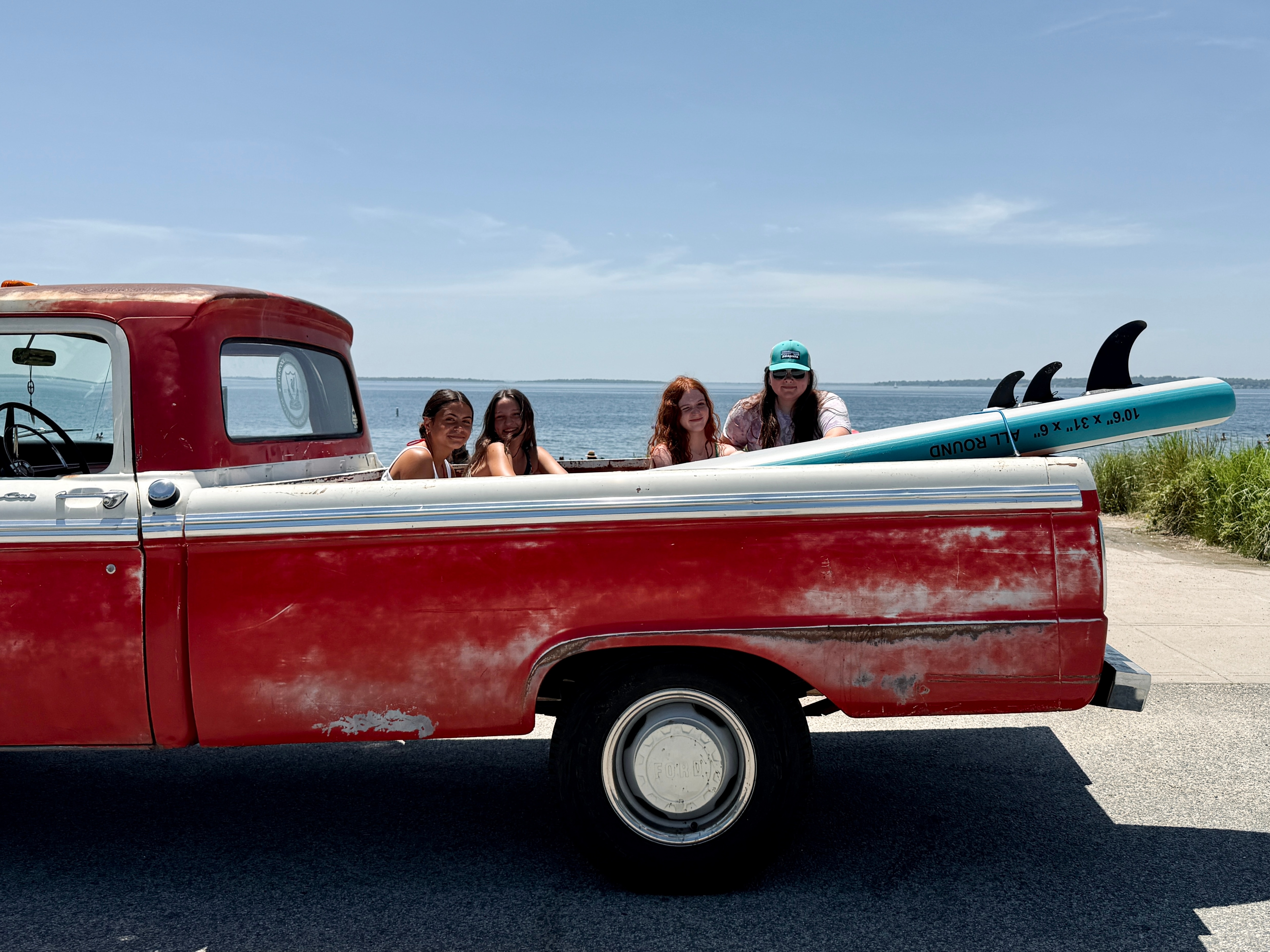 Three girls sit in the back of a vintage red truck with a surfboard. A woman leans against the side of the truck. The ocean can be seen in the background.