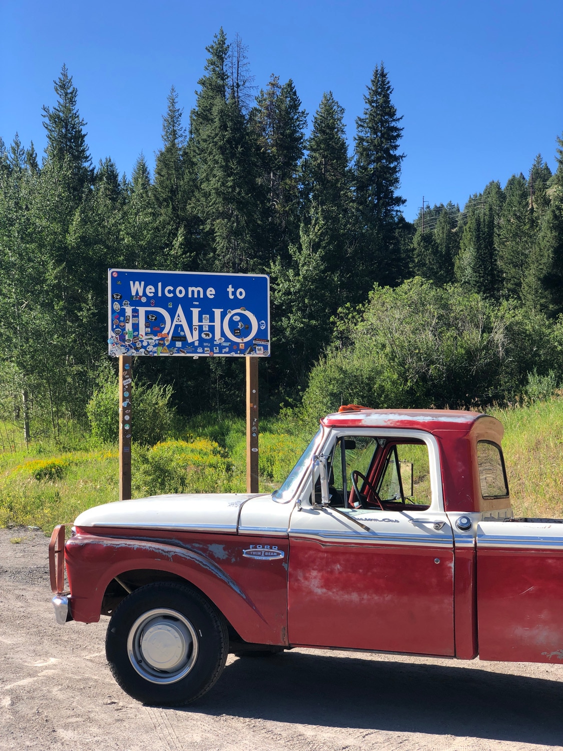 In front of a forested background, a Welcome to Idaho road sign with a vintage red truck parked in front of it.
