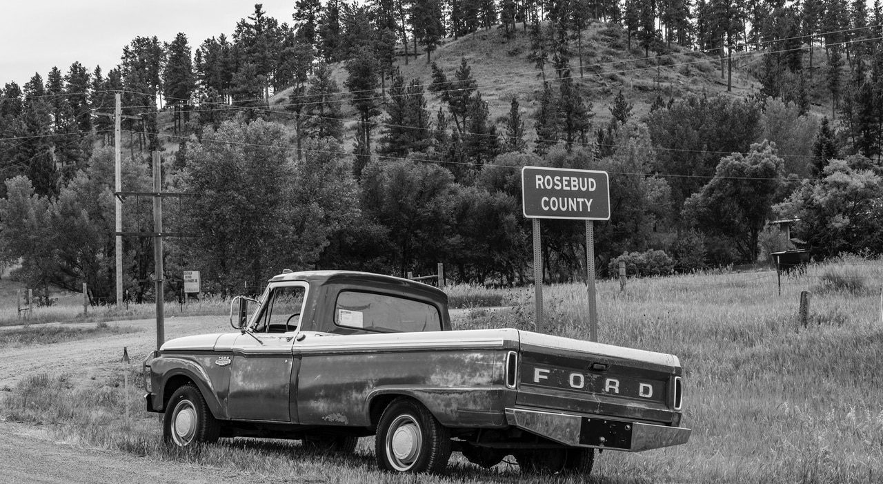 A black and white photo shows a vintage Ford truck parked next to a road sign that reads "Rosebud County" in front of a tree-covered hill.