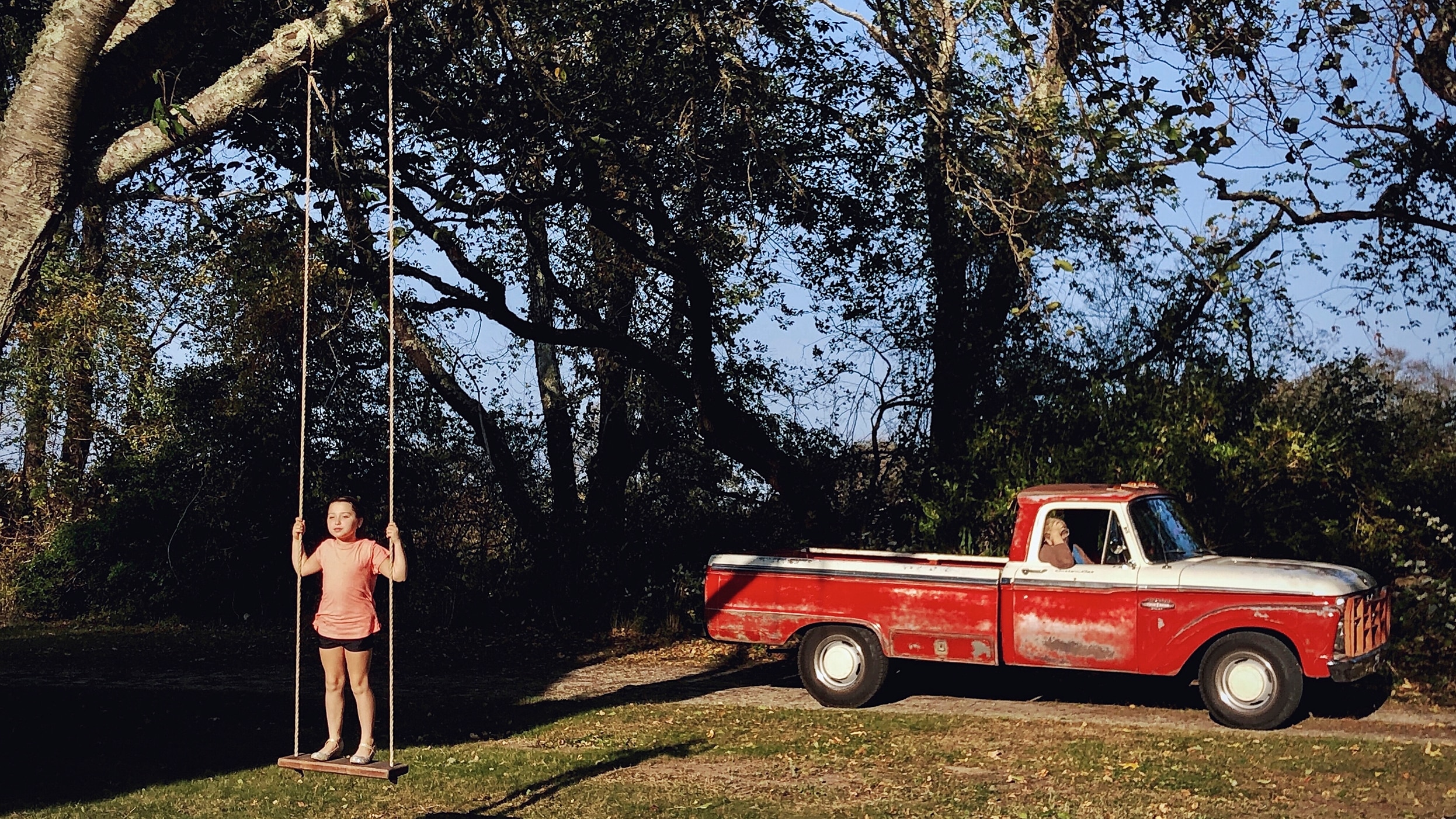 A young girl stands on a tree swing. A woman watches from the passenger seat of a vintage red truck parked next to the tree.