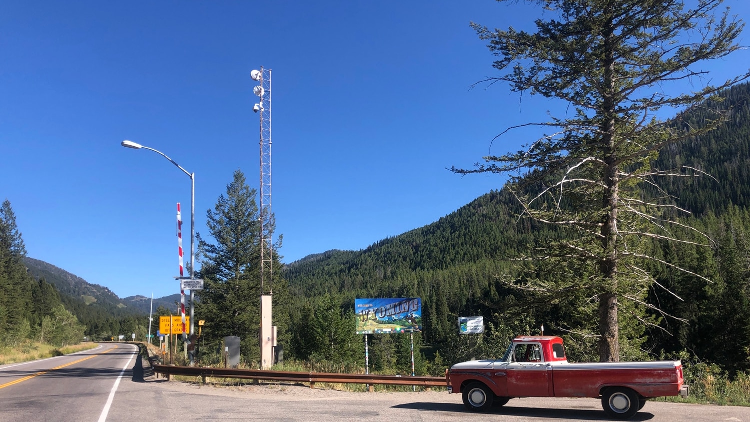 A vintage red truck parked next to the "Welcome to Wyoming" road sign in front of a mountainous forested background.