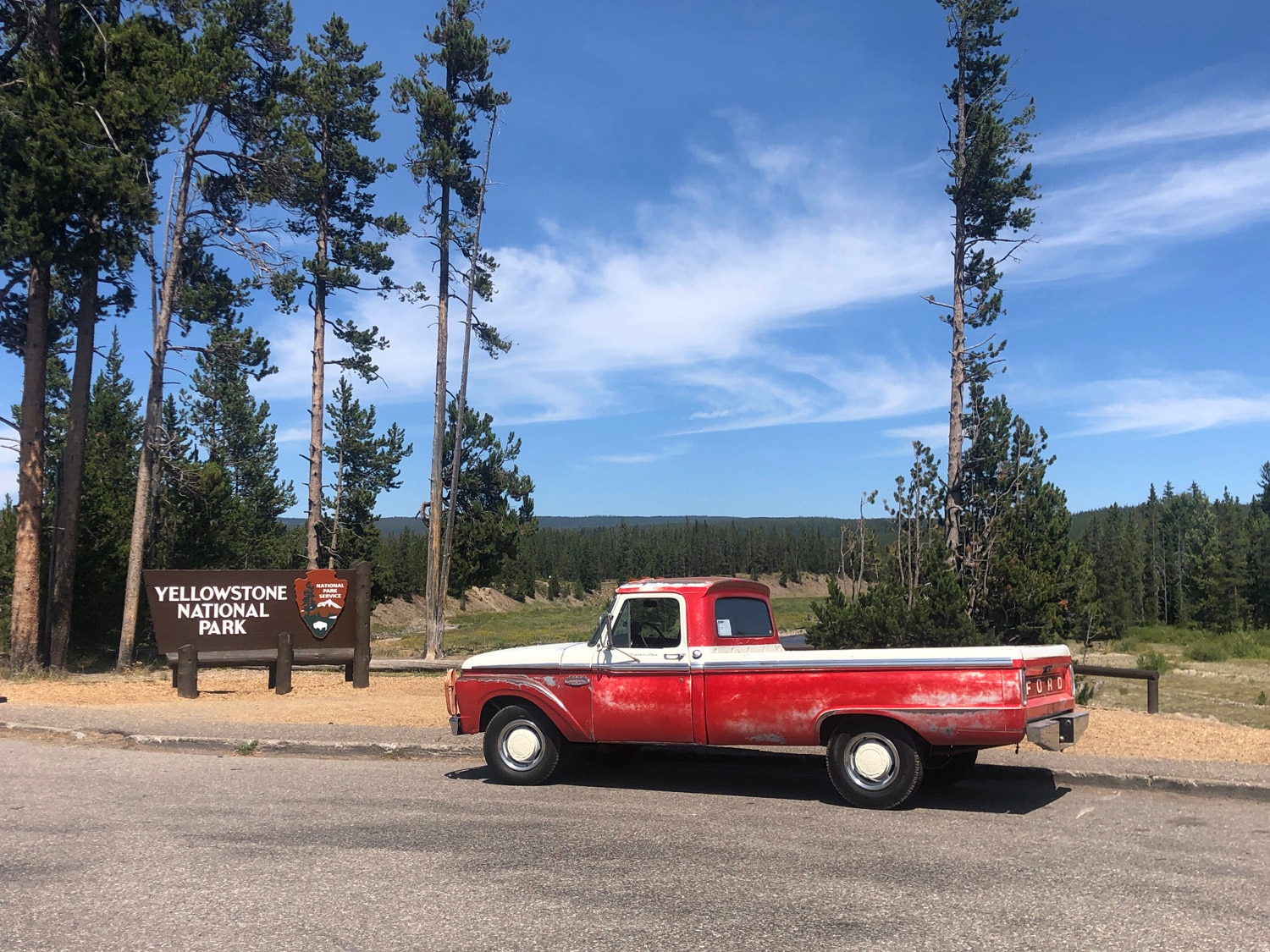 A vintage red truck parked next to a Yellowstone National Park road sign in front of a forested background.