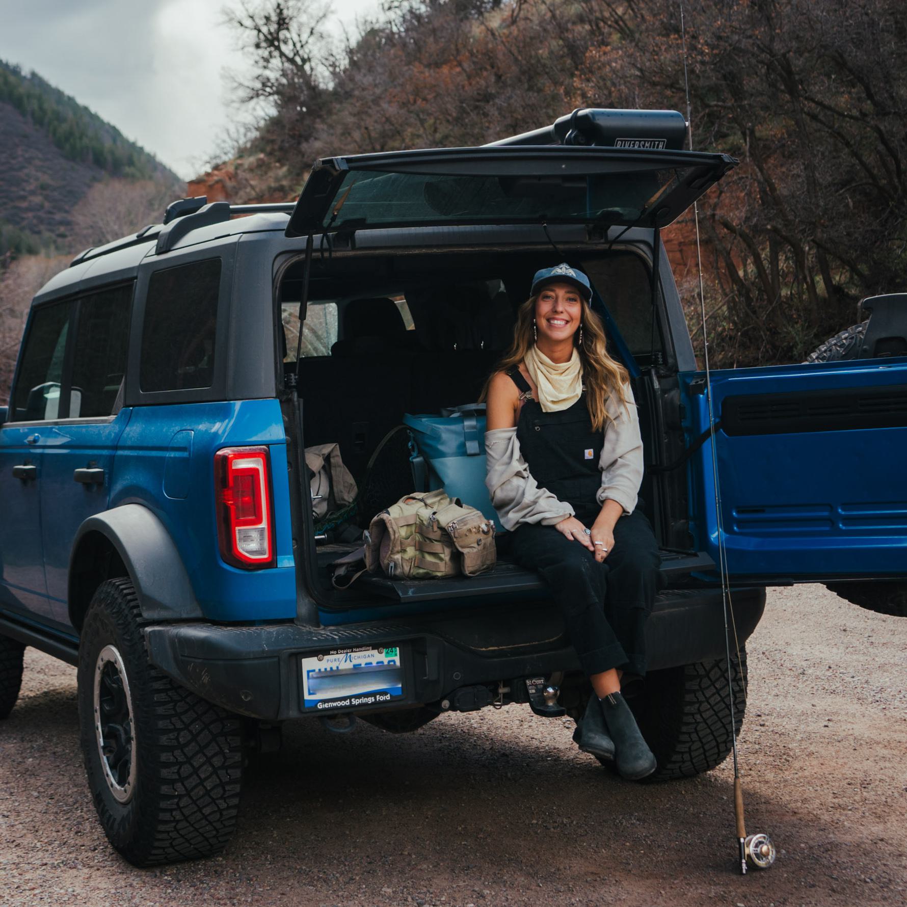 A woman in outdoor gear poses for a photo in the back of her blue Ford Bronco.