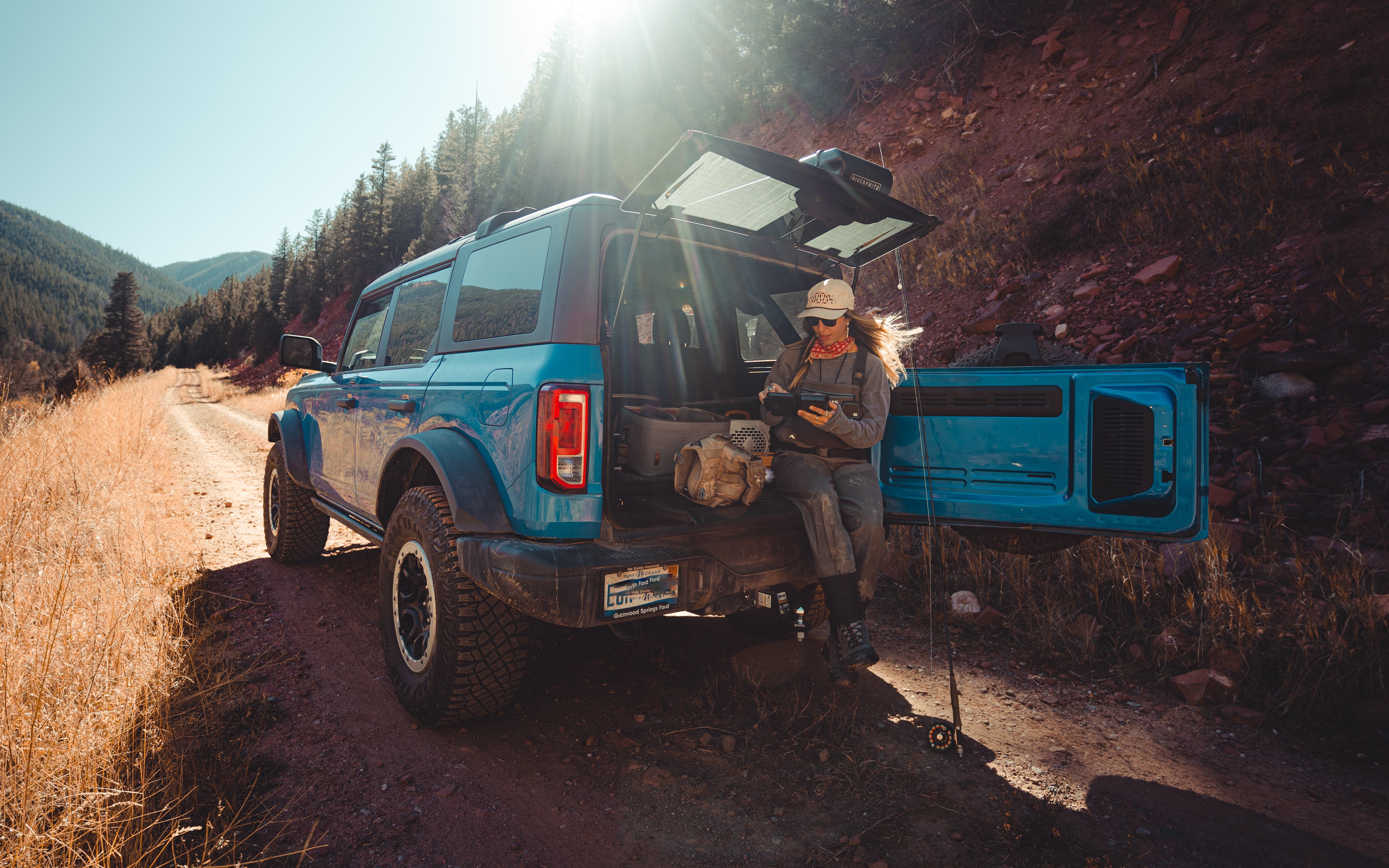 A blue Ford Bronco is parked on a dirt trail in a mountainous, forested landscape. A woman rests on the back of the Bronco  with the tailgate open.