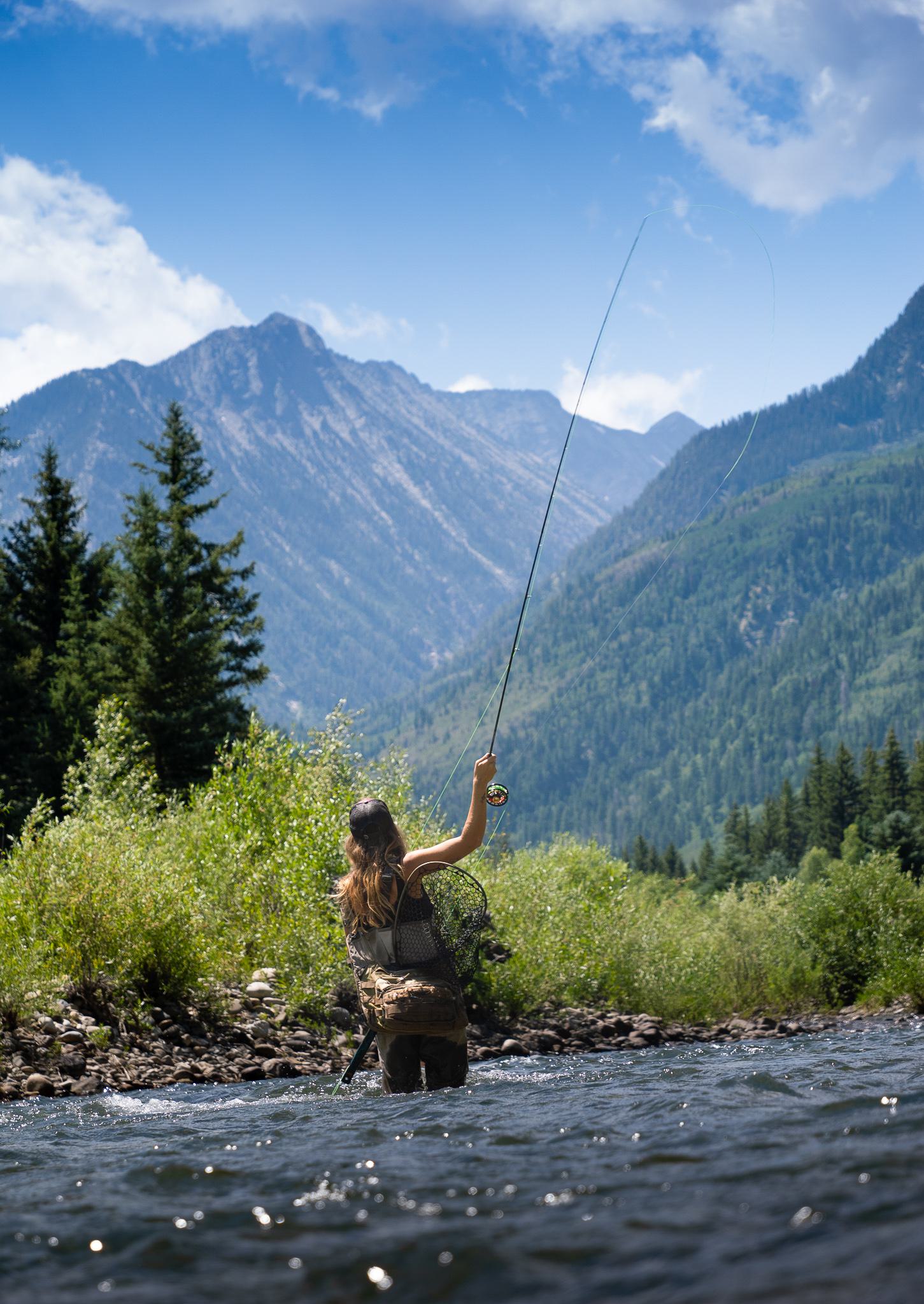 Seen from behind, a woman stands in a stream and holds a fishing pole in the air. A mountainous landscape with lush greenery surrounds her.