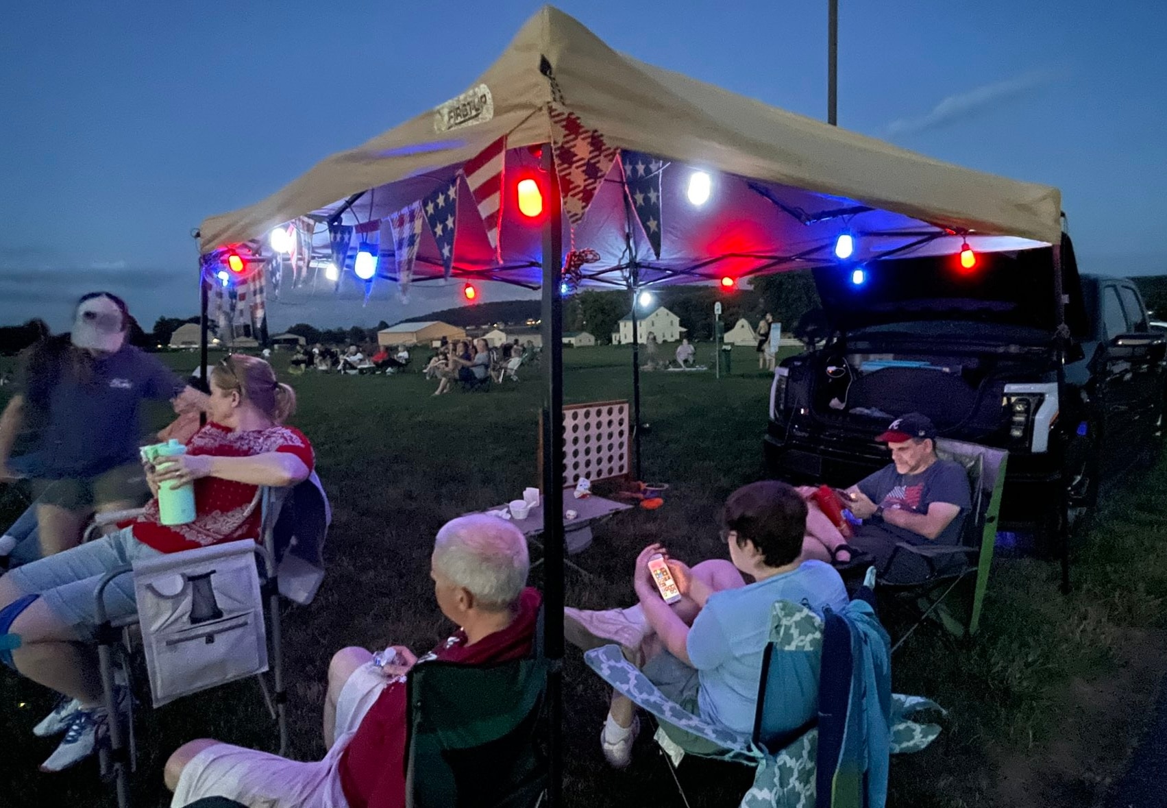 People gather around a tent decorated with red and blue lights that light up the night. Near the tent is a an electric truck with the front truck open.