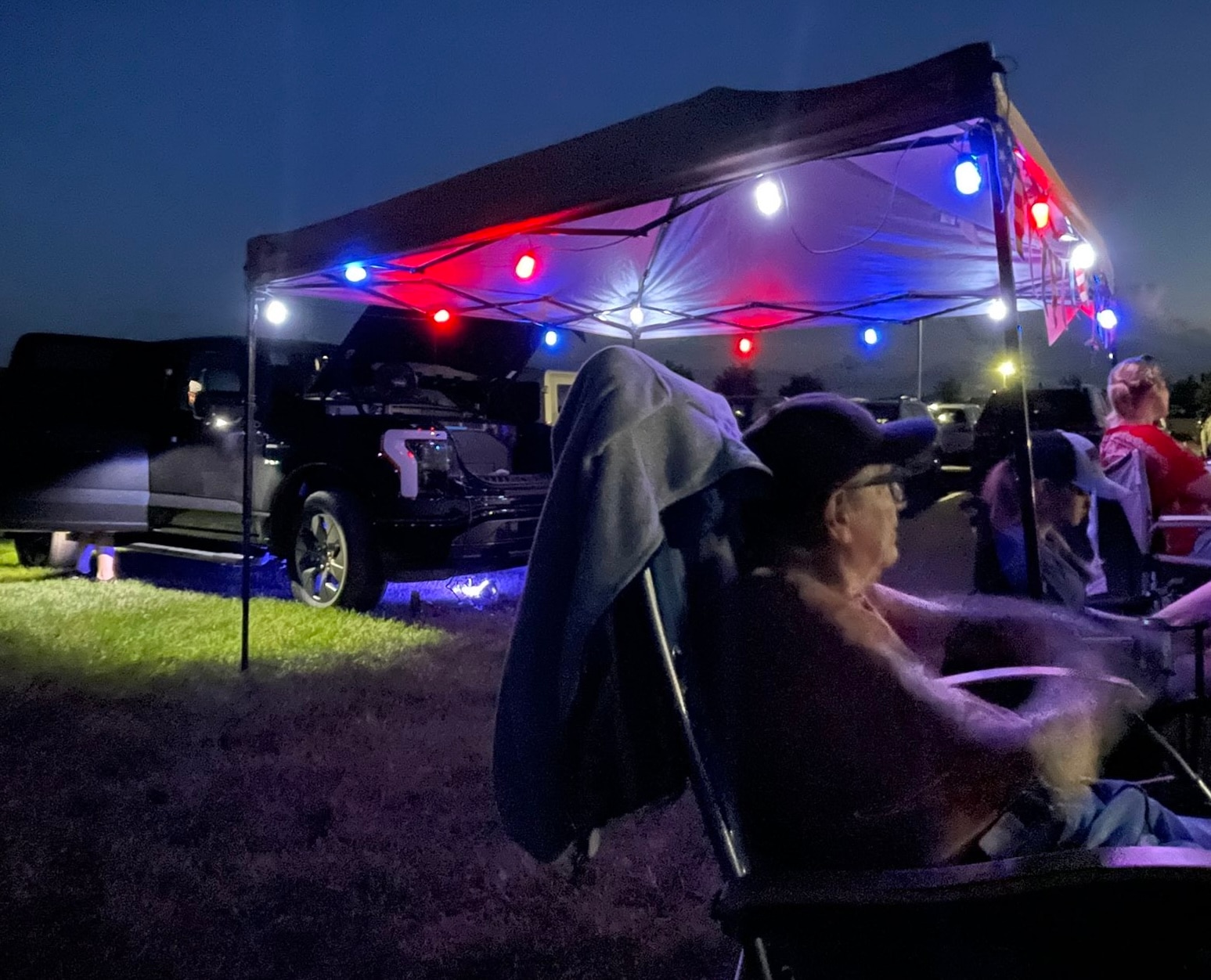 People gather around a tent decorated with red and blue lights that light up the night. Near the tent is a an electric truck with the front truck open.