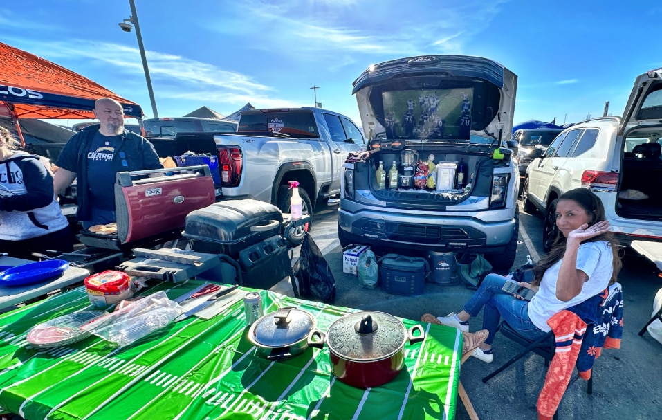 A table with food sits in front of the front trunk of an electric vehicle, which holds drinks and a tv