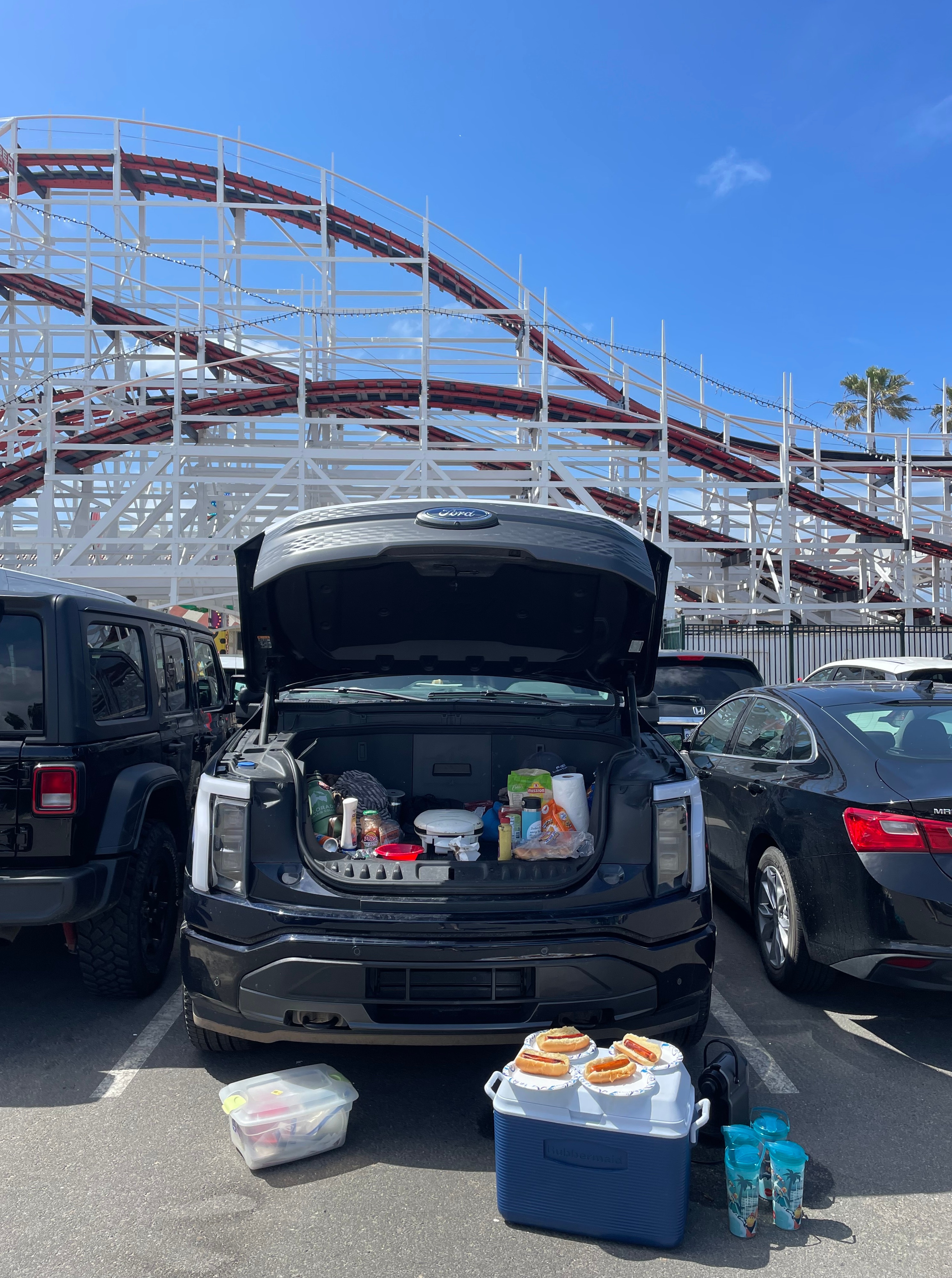 A cooler with hot dogs on it sits in front of the open front trunk of an electric vehicle, which holds the condiments and other items