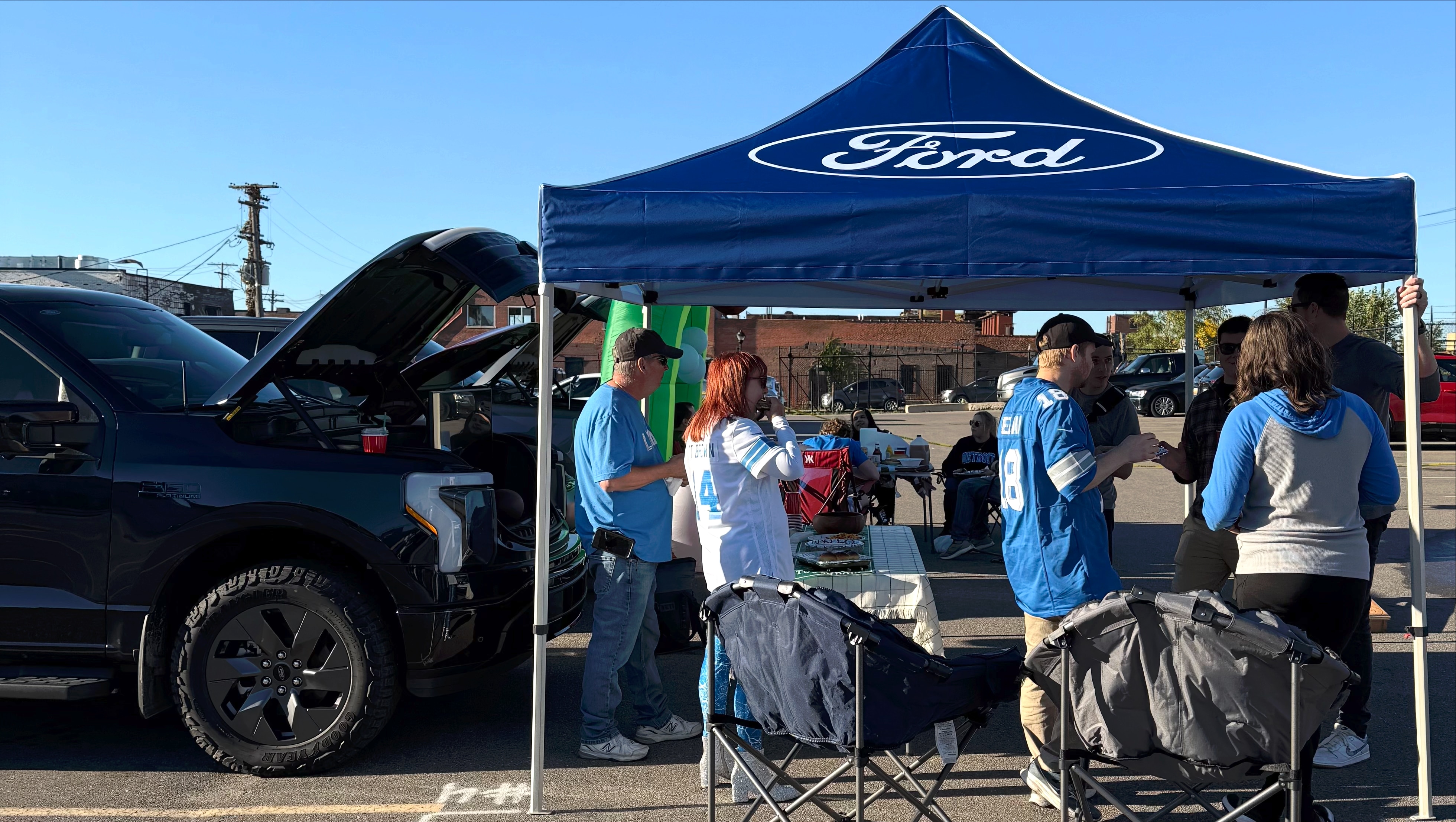 People gather under a tent with the word "Ford" on it in front of the open front trunk of an electric vehicle