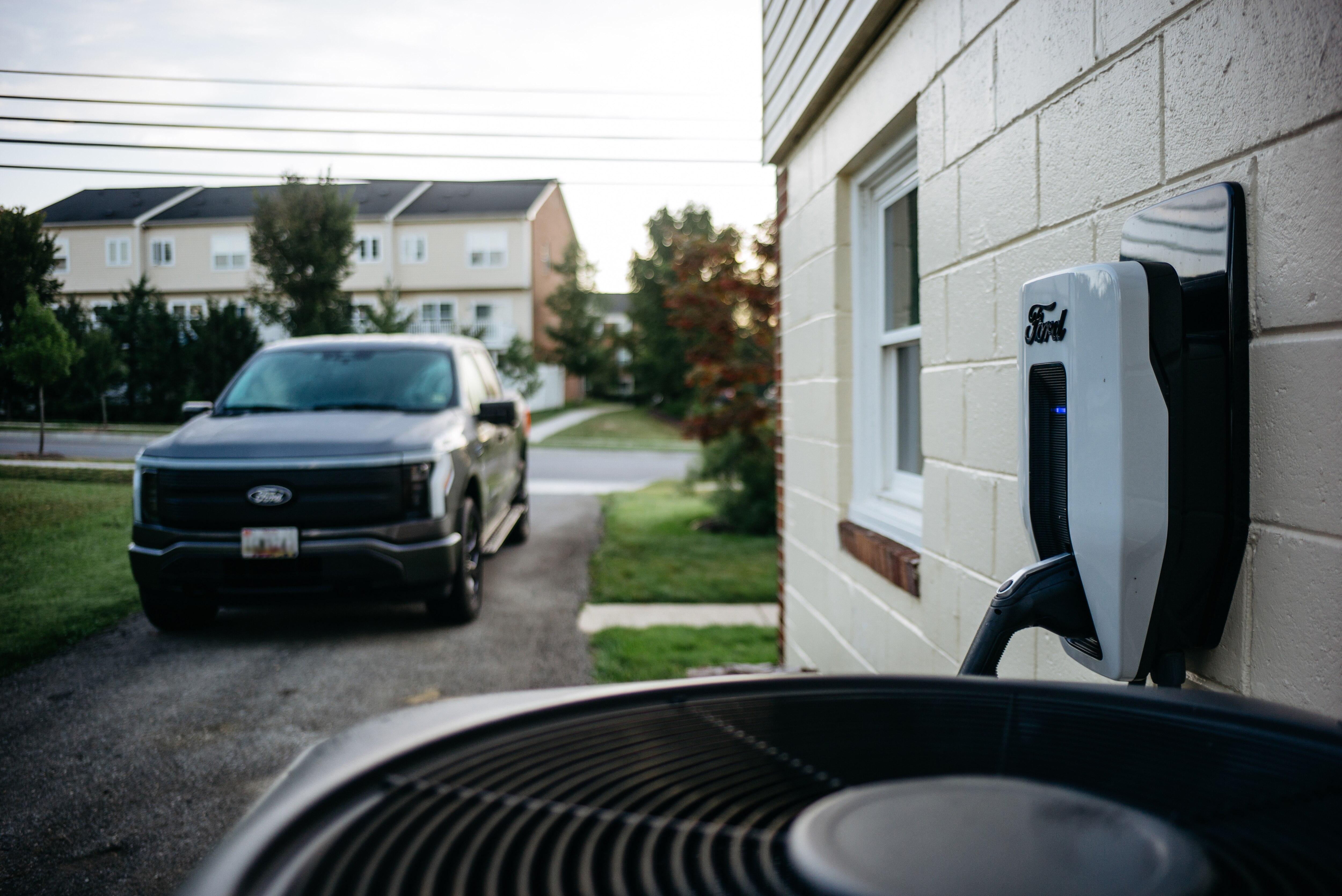 A white Ford charger is on the side of a house with cream painted bricks. In the background, a grey Ford F-150 Lightning is parked in the driveway.