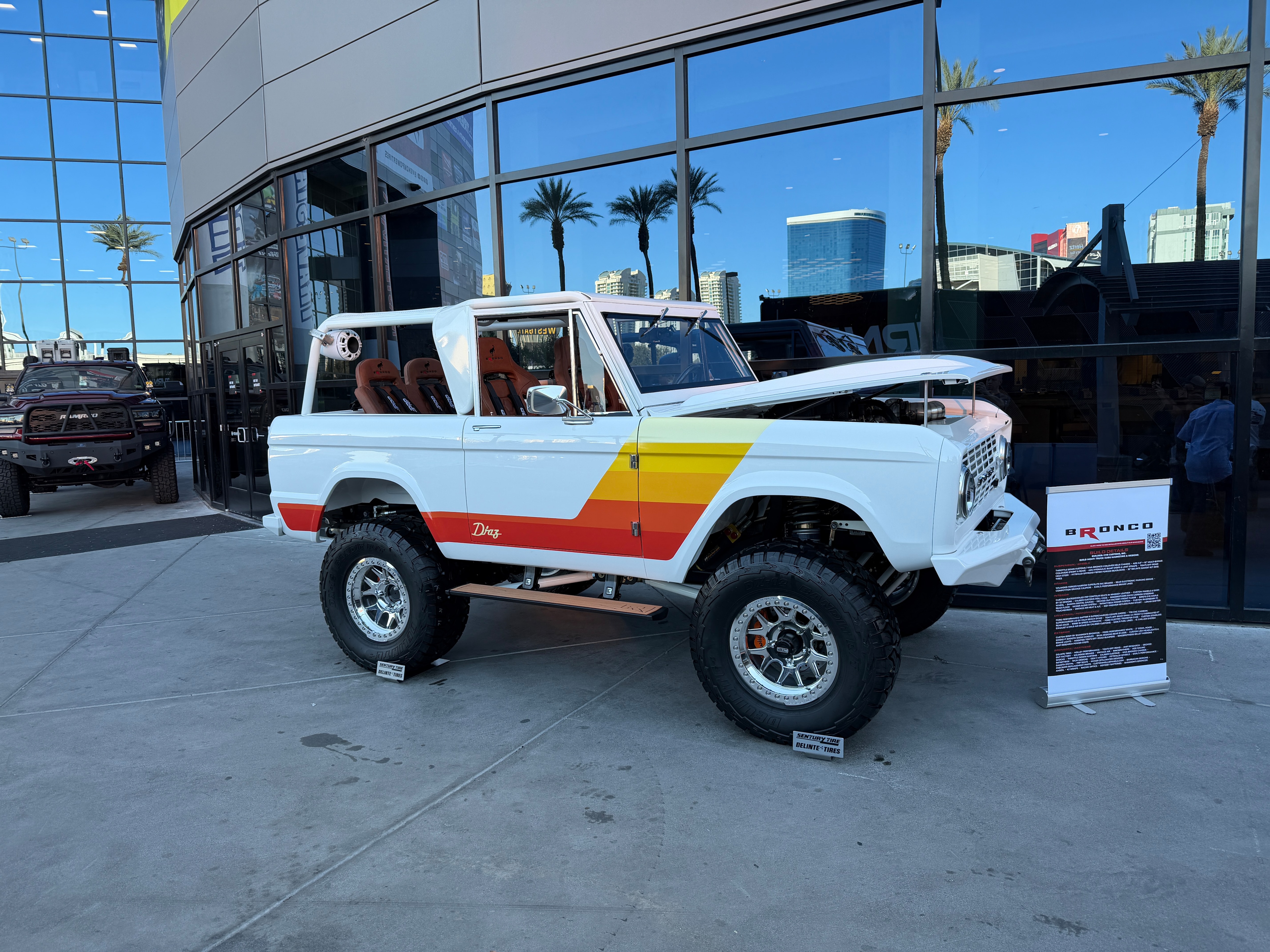 A vintage-looking white bronco with orange and yellow side-decals displayed at a car show