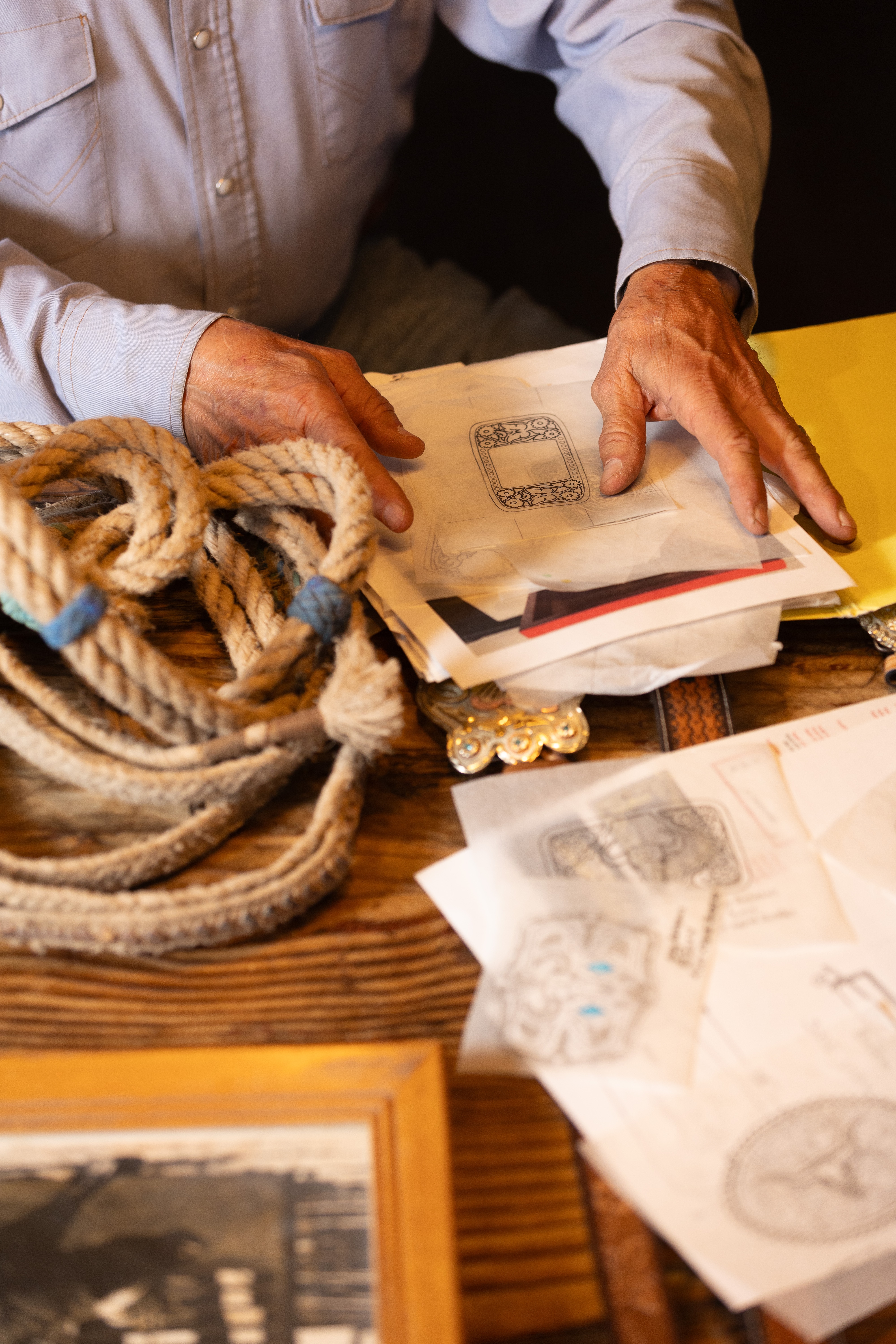 A man holds a sketch on transparent paper, surrounded by rope and other tools he uses to craft items such as belt buckles.
