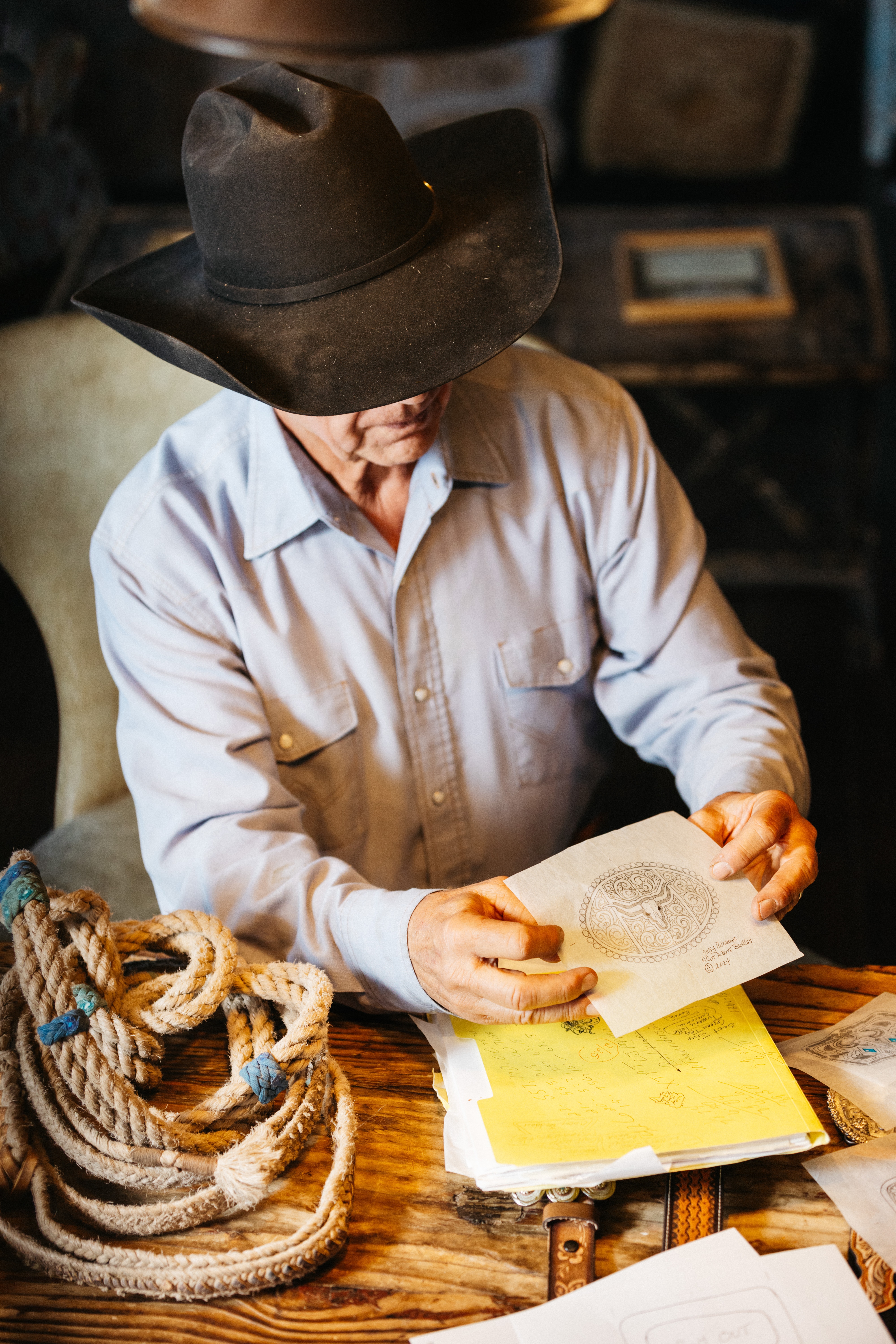 A man holds a piece of thin, transparent paper with a drawing on it in his workshop. He is dressed in Western attire.