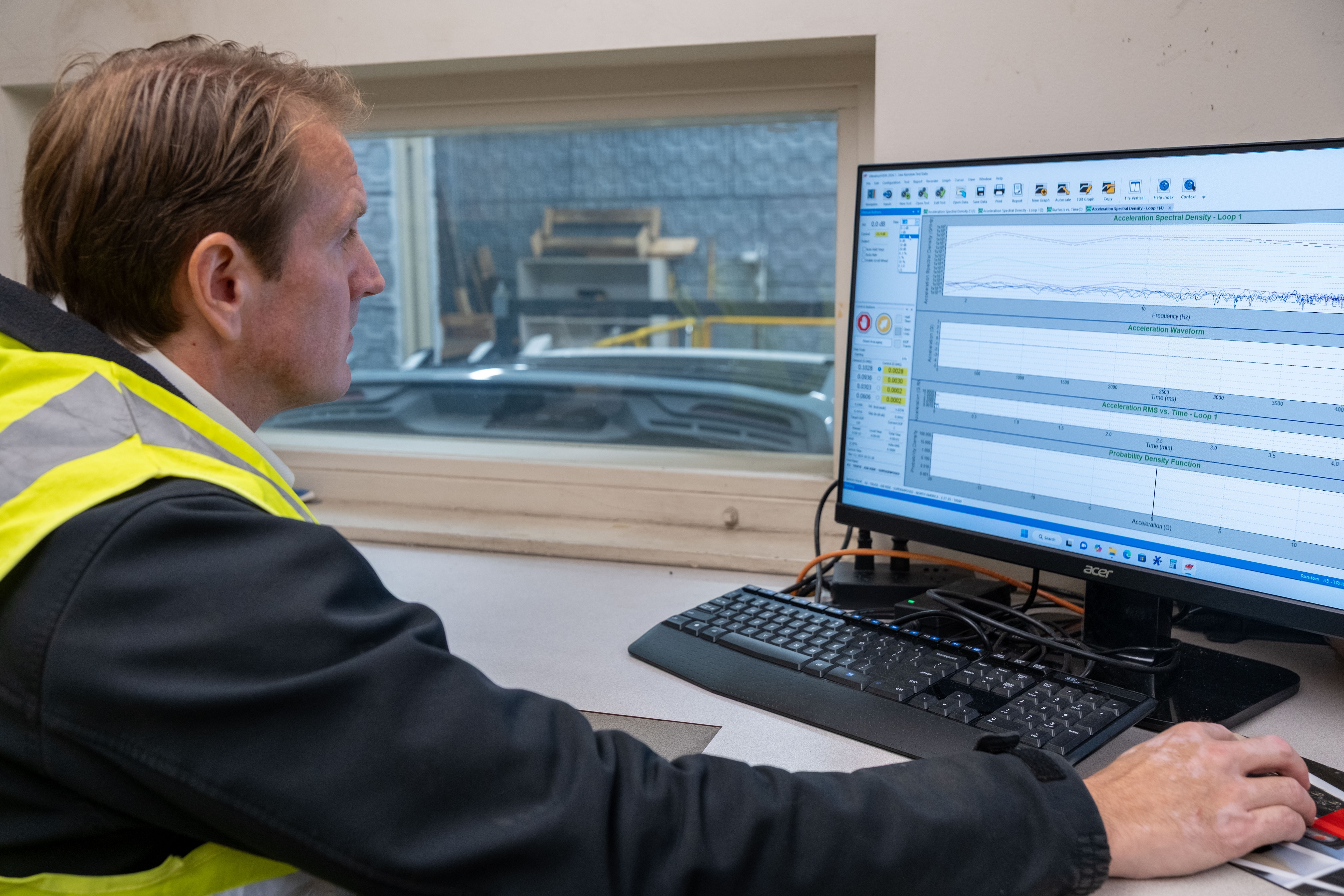 A man in a yellow high-visibility vest looks at a computer monitor near a window overlooking manufacturing testing equipment.