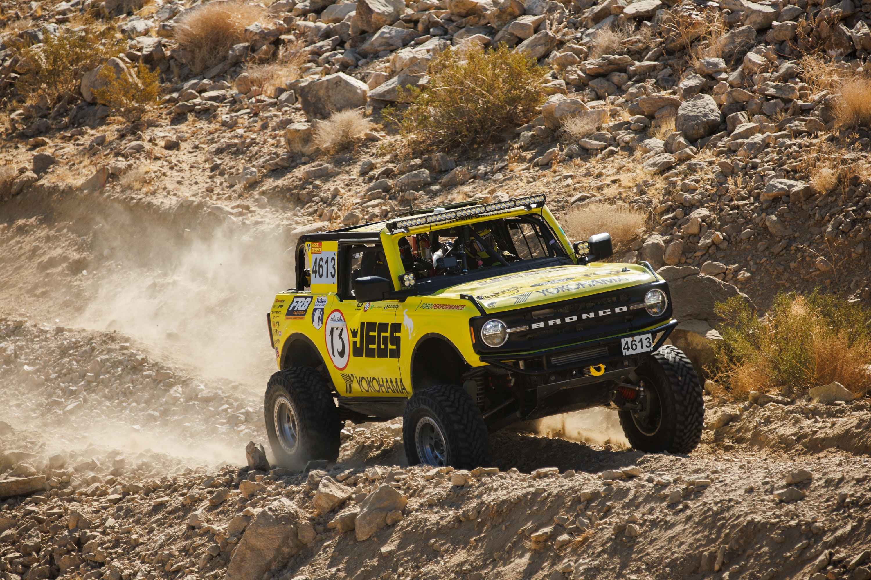 A yellow Bronco drives off road on rocky terrain.