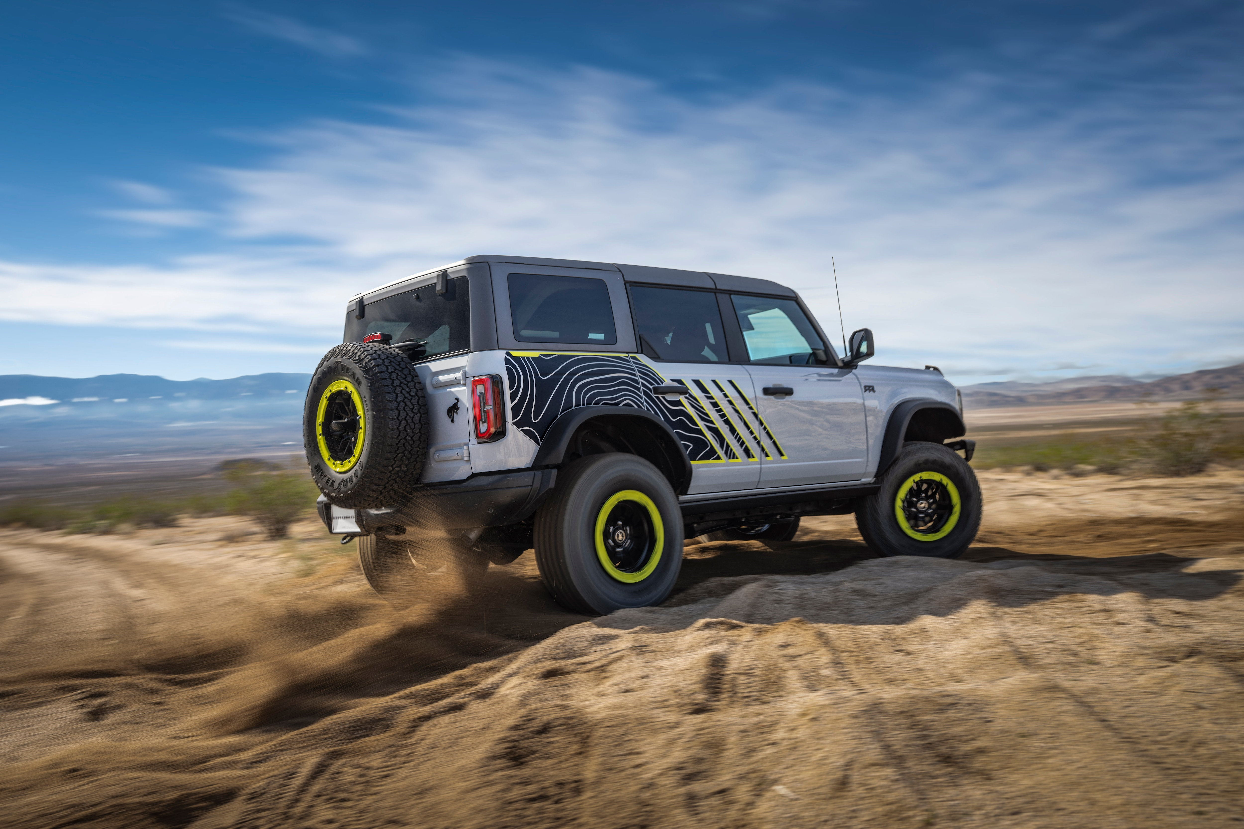 Side view of the Bronco RTR driving through sand with a blue slide and whispy clouds overhead. The vehicle is silver with lime green highlights on the wheels and side.
