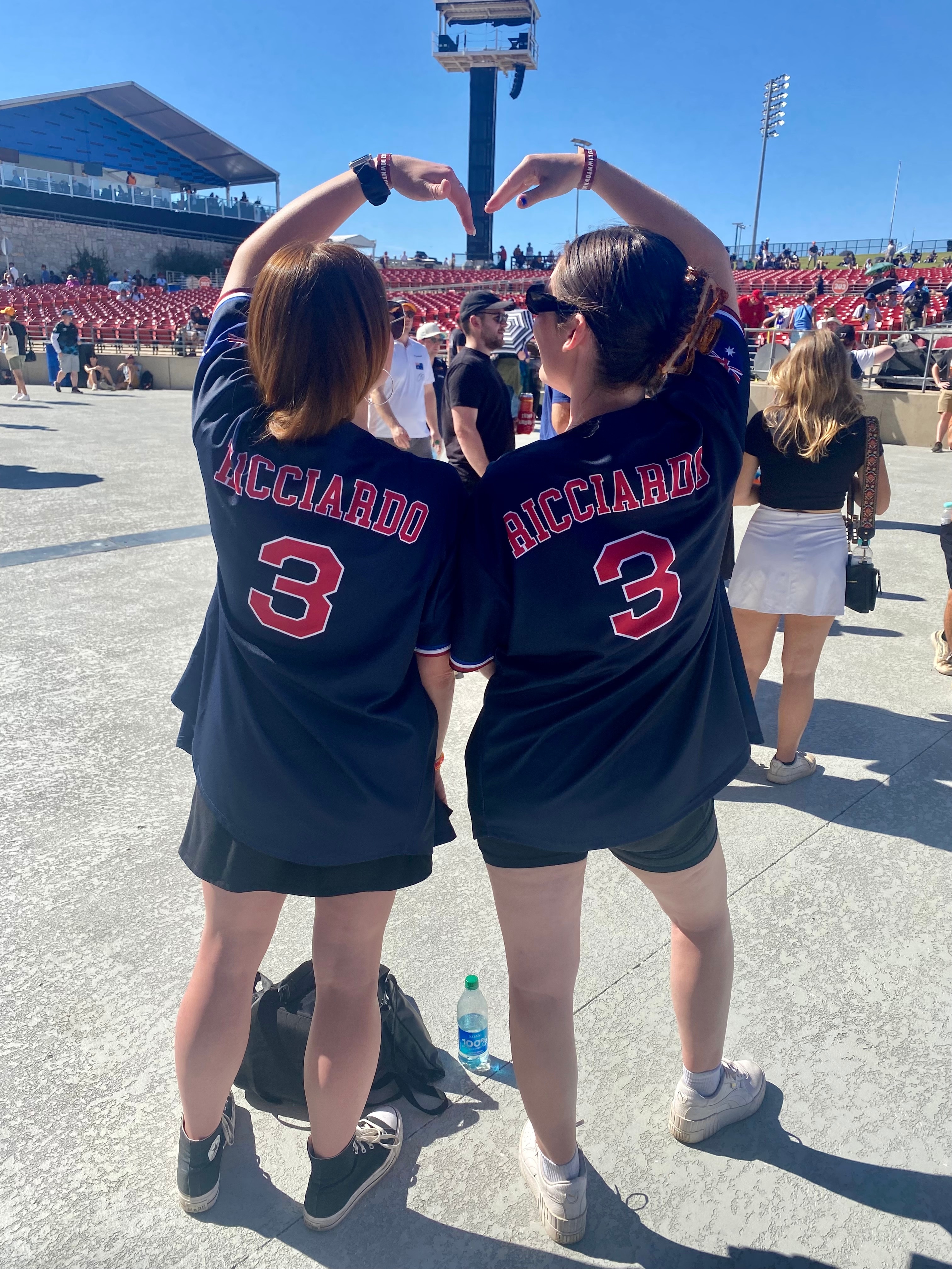 Viewed from the back, two women in t shirts that read "Ricciardo 3" on the back make a heart shape with their arms in front of a race track.