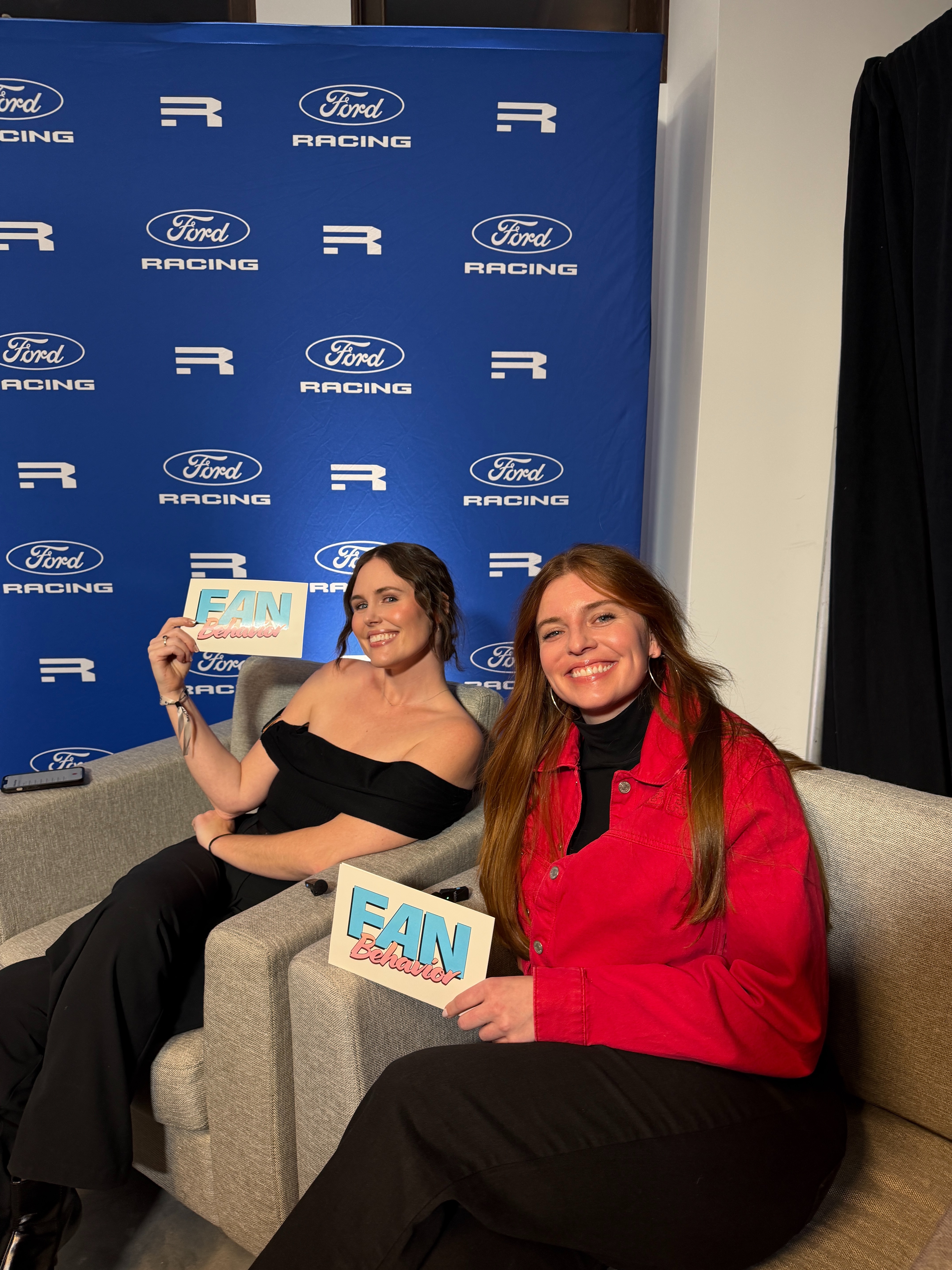 Two women seated on a stage in front of a Ford Racing backdrop hold signs that say "Fan Behavior."