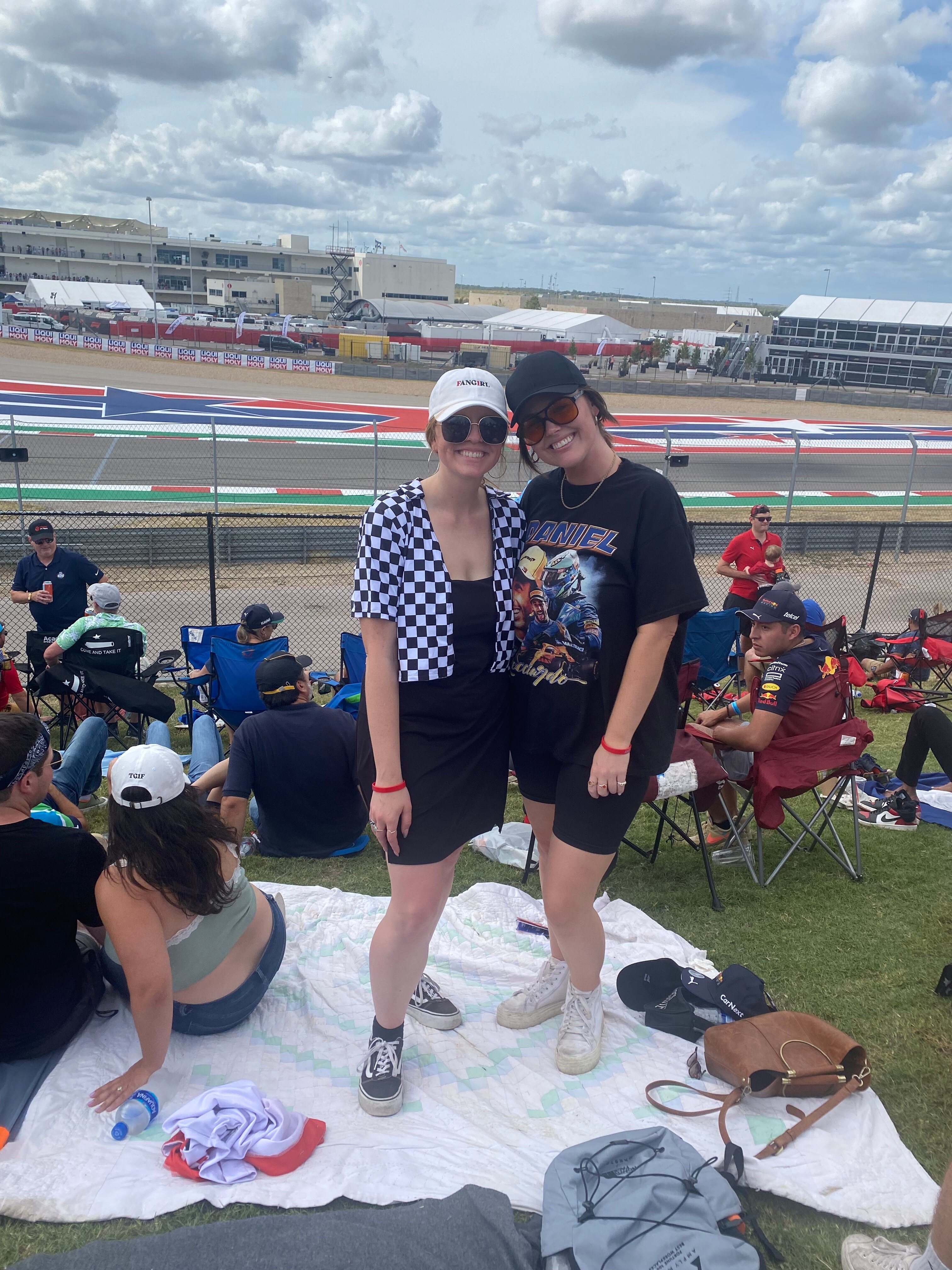 Two women stand on a blanket, posing for a photo in front of a race track.