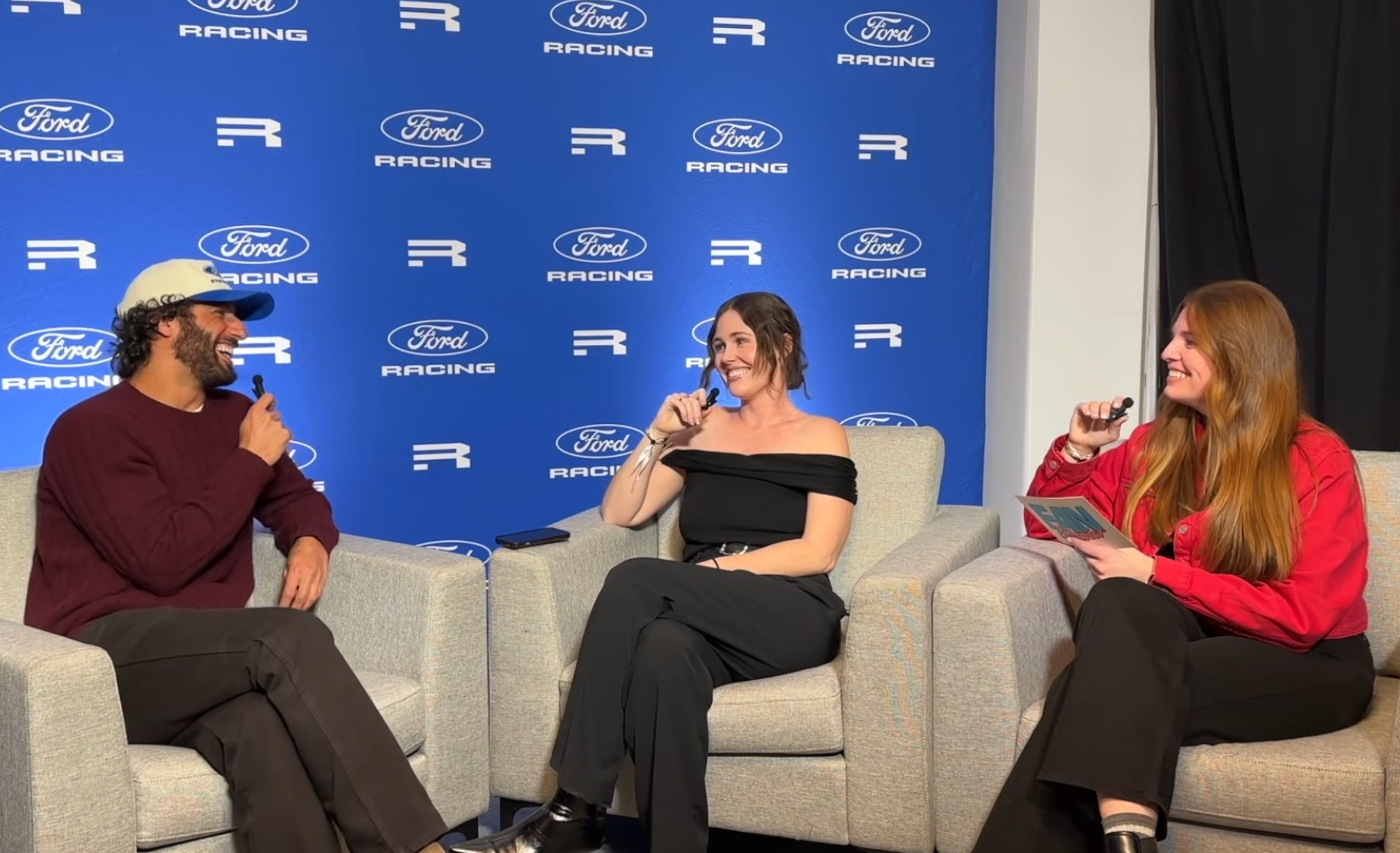 Two women interview Daniel Riccardio on stage with a Ford Racing blue backdrop.