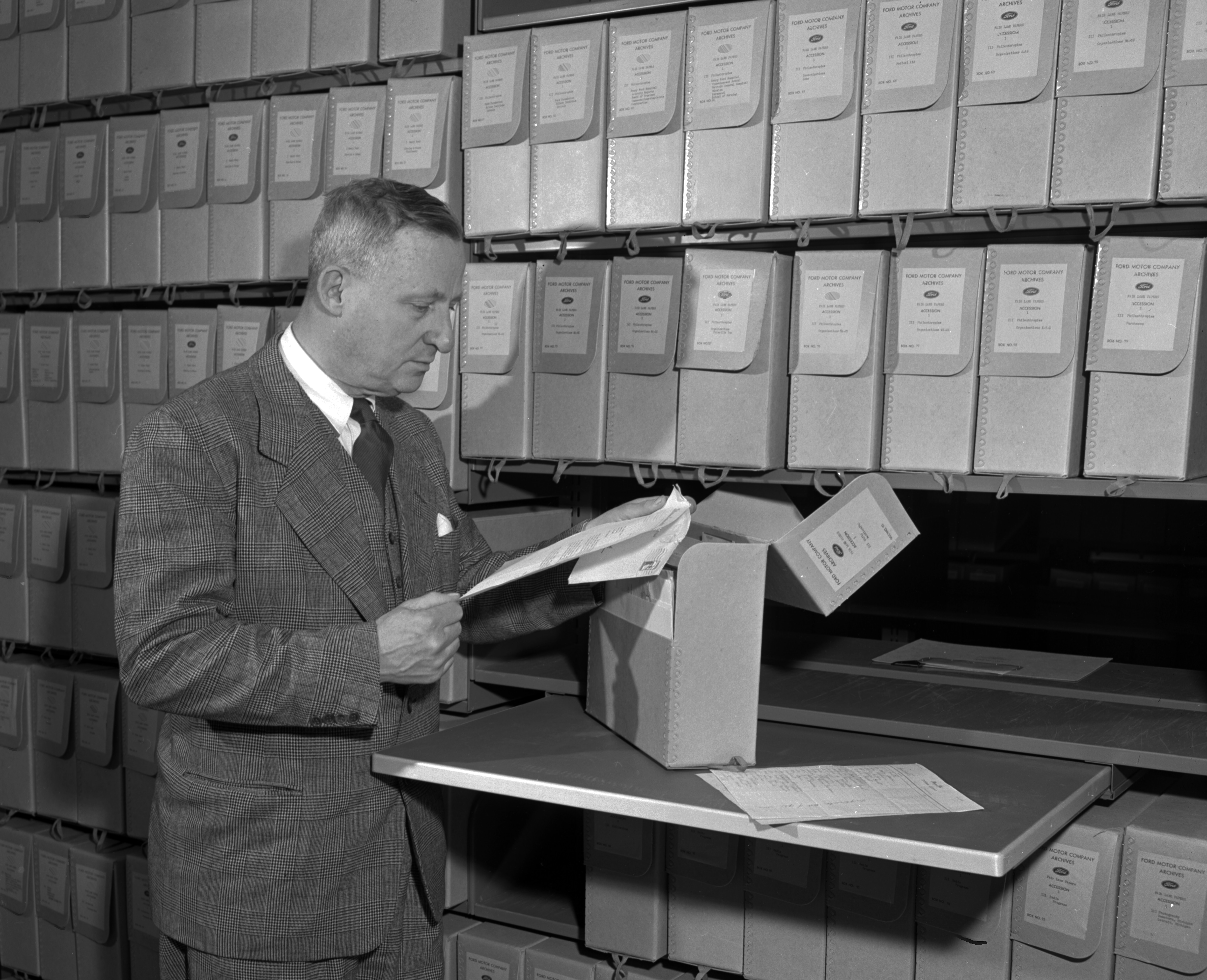 A black and white photo shows a man reading files that he has pulled out of a box in the archives.