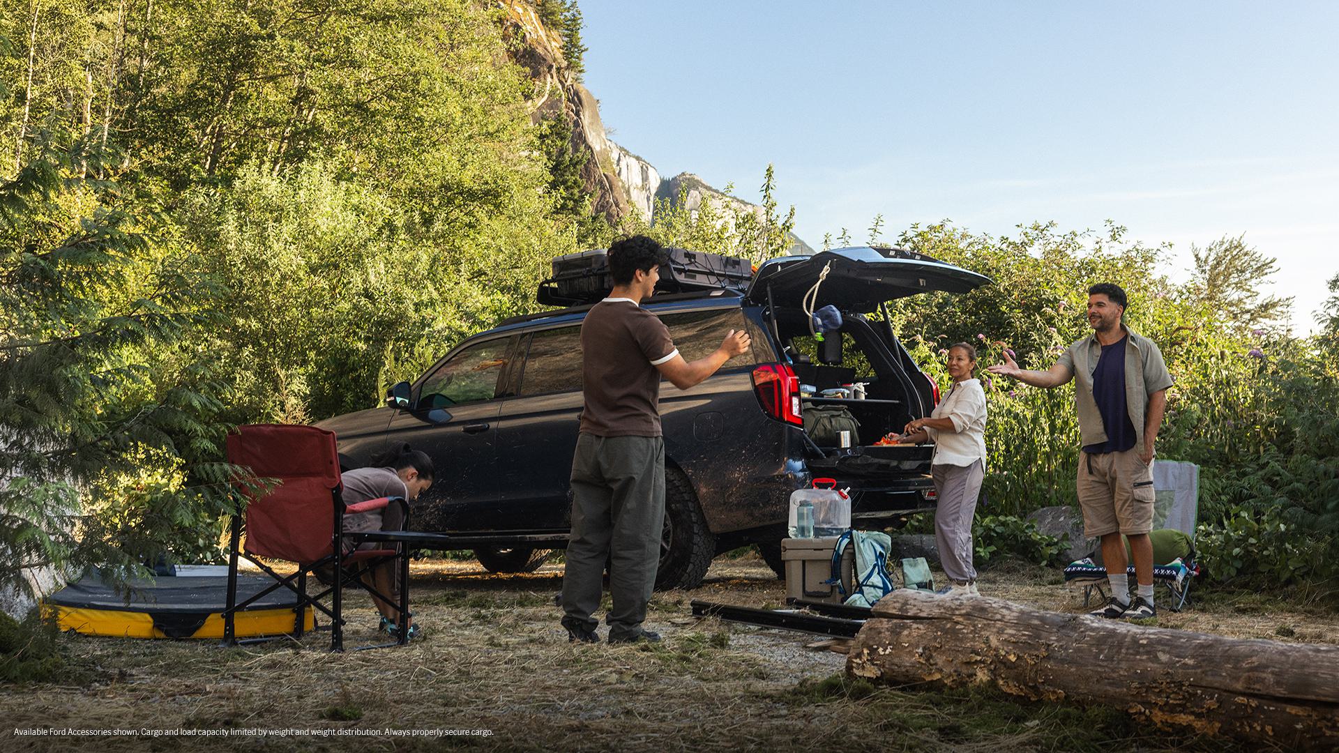 A family hangs out at a campsite in the woods, using the open back of their vehicle as a table and storage.