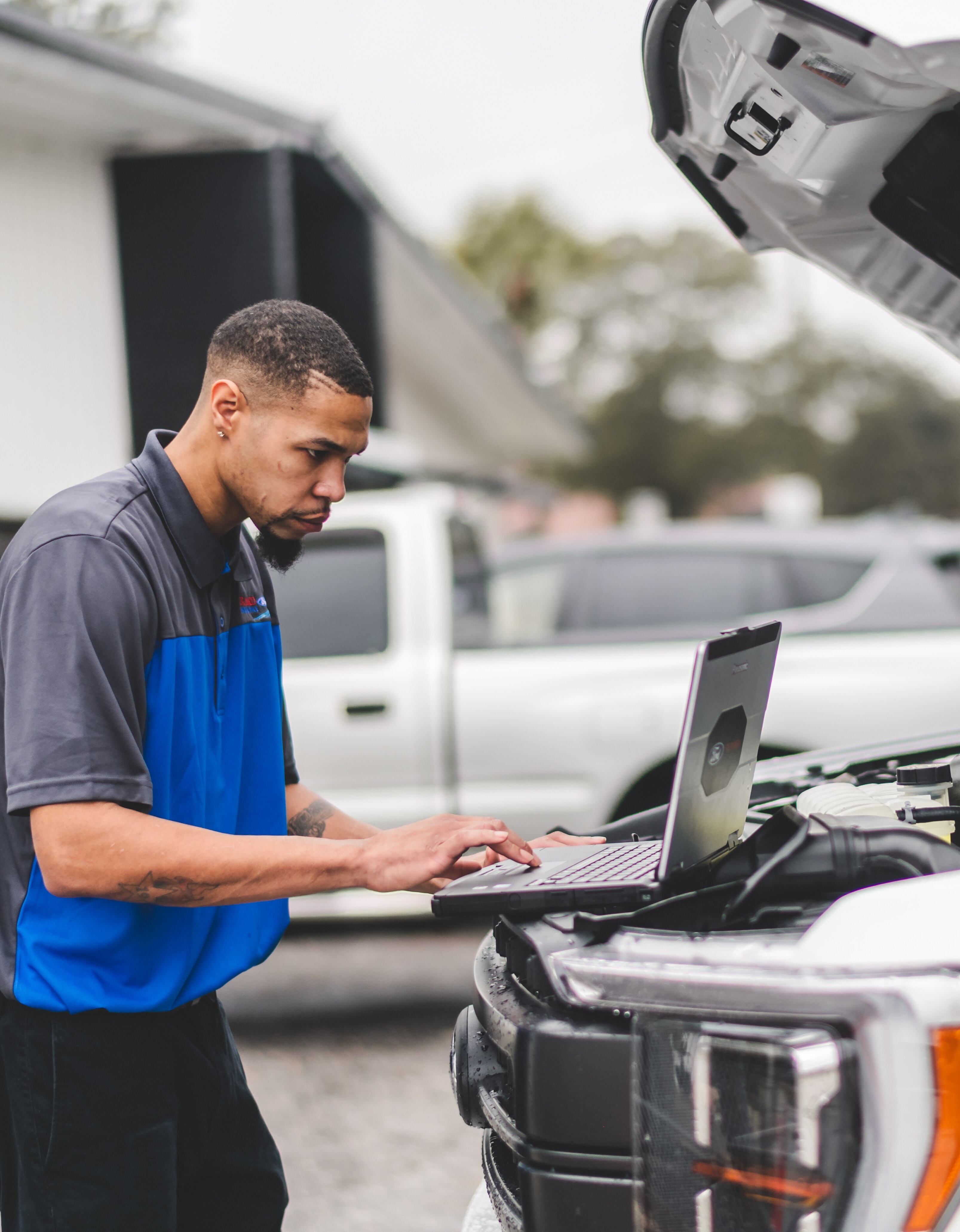 A man looks at a laptop sitting under the open hood of a truck.